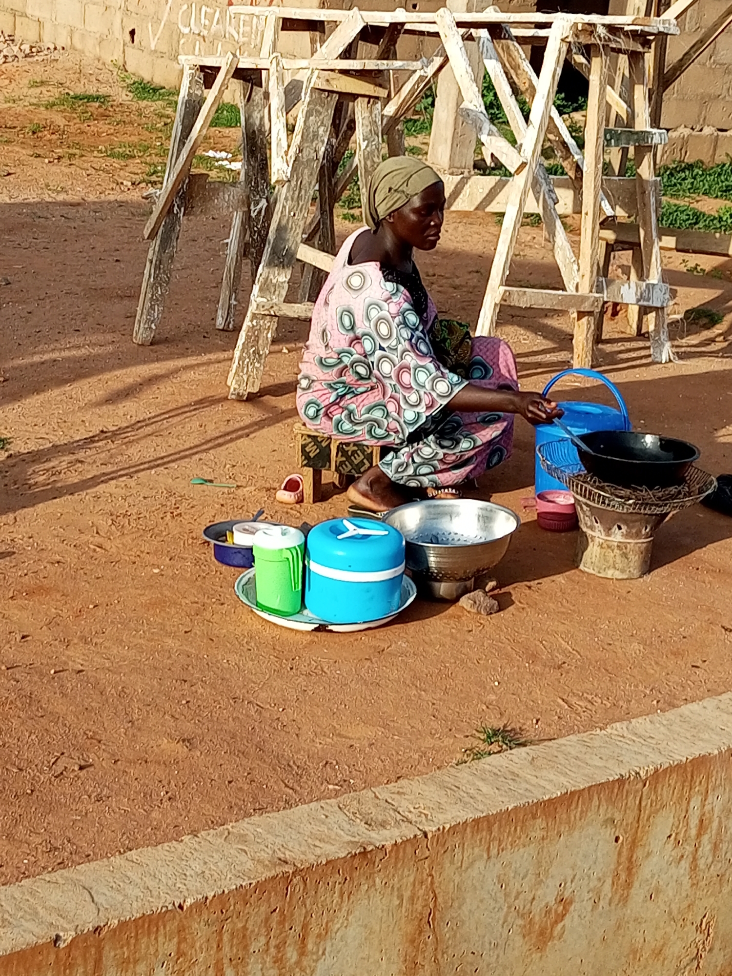 In Tempure, Nigeria, a woman is seated on a wooden stool in an outdoor setting. She is wearing a headscarf and a colorful dress. In front of her, on the ground, are several items arranged on a tray: a green container, a blue circular container, and a metal bowl. To the right of these items, a metal stove is visible, with a black pot placed on top. A blue plastic bucket stands next to the stove. The background consists of a rough wooden structure, a wall with some graffiti, and patches of grass. The ground is covered in reddish-brown soil, and shadows are cast by the structures and objects, suggesting it is daytime.