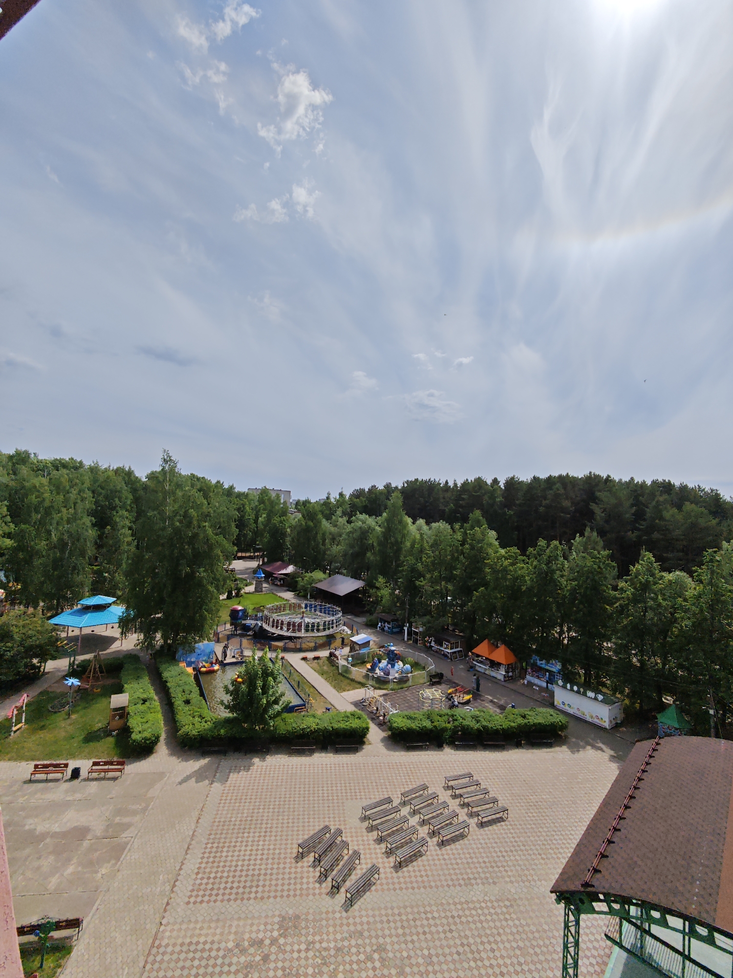The image shows an aerial view of a park in the city of Chaikovsky, Russia. The park includes several rides and attractions, including a carousel, a small pond with boats, and a playground. There are also several benches and picnic tables in the park. The park is surrounded by trees and greenery. The sky is partly cloudy.