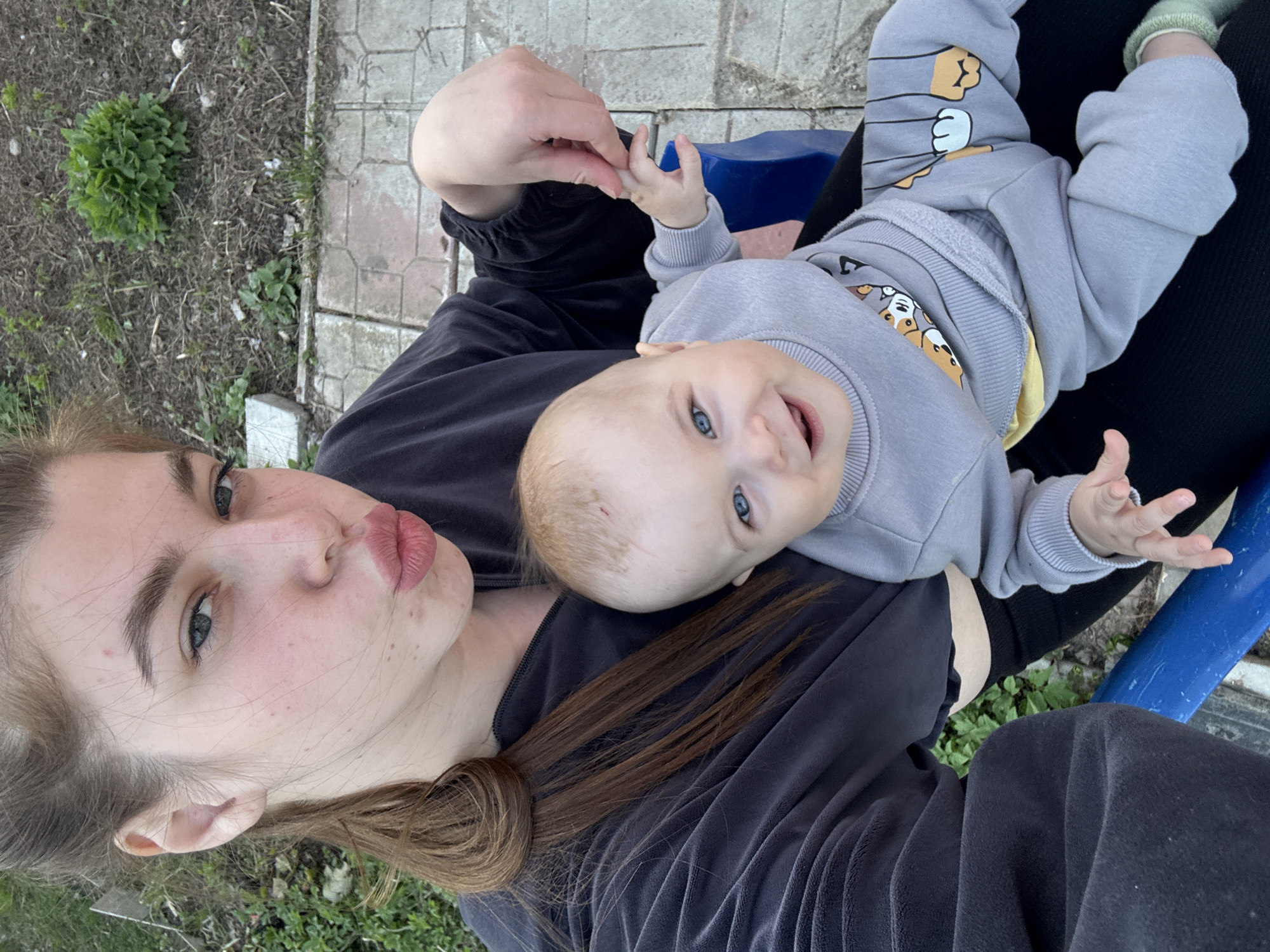 A young woman and a baby are outdoors. The woman, with brown hair tied back, is wearing a dark grey, velvety tracksuit. She has light skin, freckles, and is pouting slightly towards the camera. The baby is lying on her lap, wearing a light grey tracksuit with cartoon designs. The baby has blue eyes and is smiling. The background shows grass and some paving. It's daytime. The location is most likely a park or garden, but there is no evidence to suggest this is located in Мещёры, Russia.