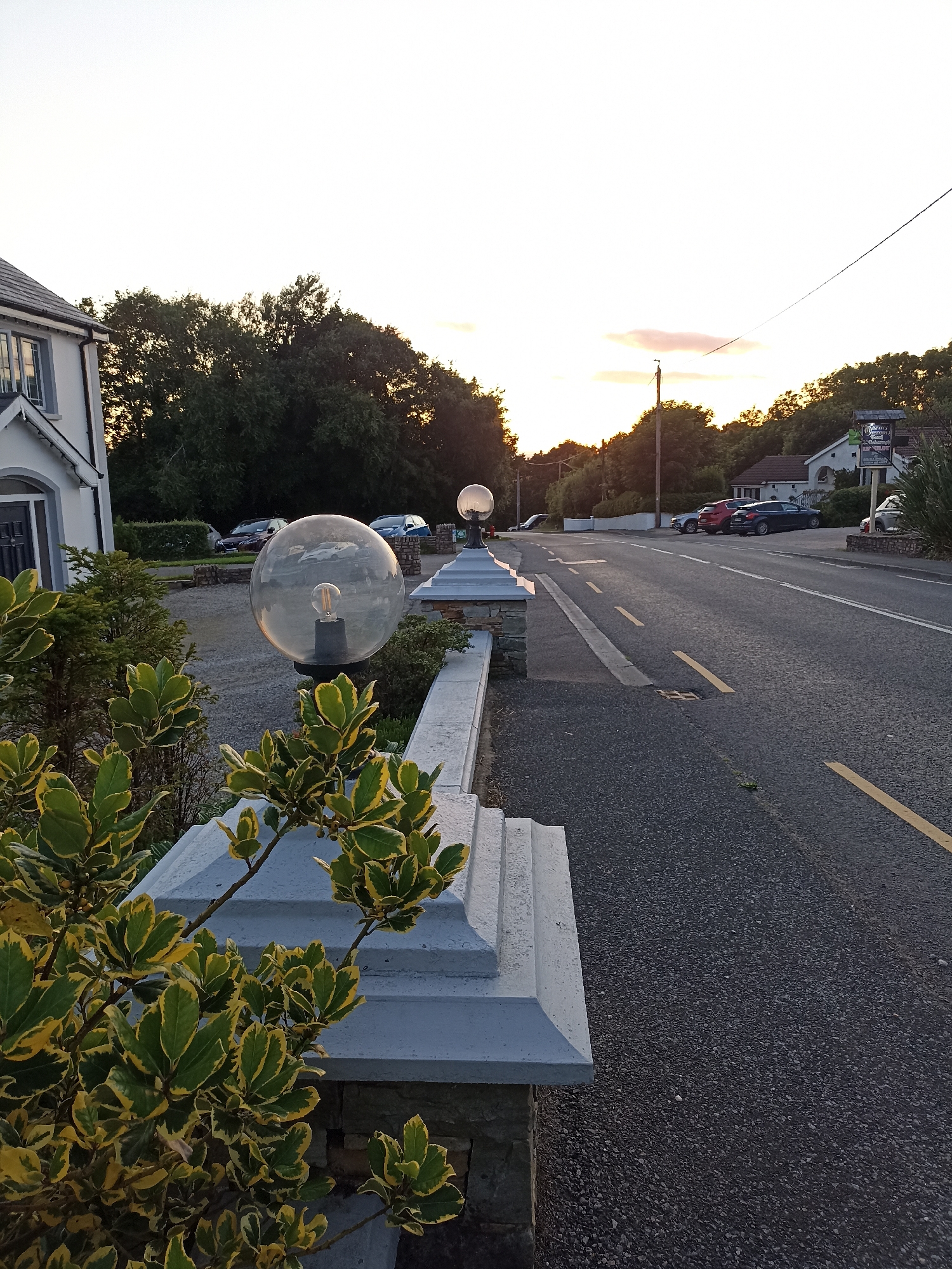 The image depicts a street scene in Annagry, Ireland, at dusk. In the foreground, a variegated shrub with green and yellow leaves is positioned in front of a stone and concrete pillar, which supports a large, clear glass globe lamp with an illuminated filament. To the left, a portion of a white building with grey trim and a dark door is visible. Beyond the building, a dense line of dark green trees forms the background.
The street, paved with asphalt, stretches from the foreground into the distance, marked by yellow lines. Several cars are parked along the side of the road. In the middle ground, another similar globe lamp on a pillar is visible. The sky above is a pale, washed-out blue with a hint of pink and orange near the horizon, suggesting the sun is setting. A single utility pole with a wire stretching across the frame is also present. A sign for "Mary G's" is mounted on a pole near the right side of the road. The overall impression is of a quiet, semi-rural setting as evening approaches.
