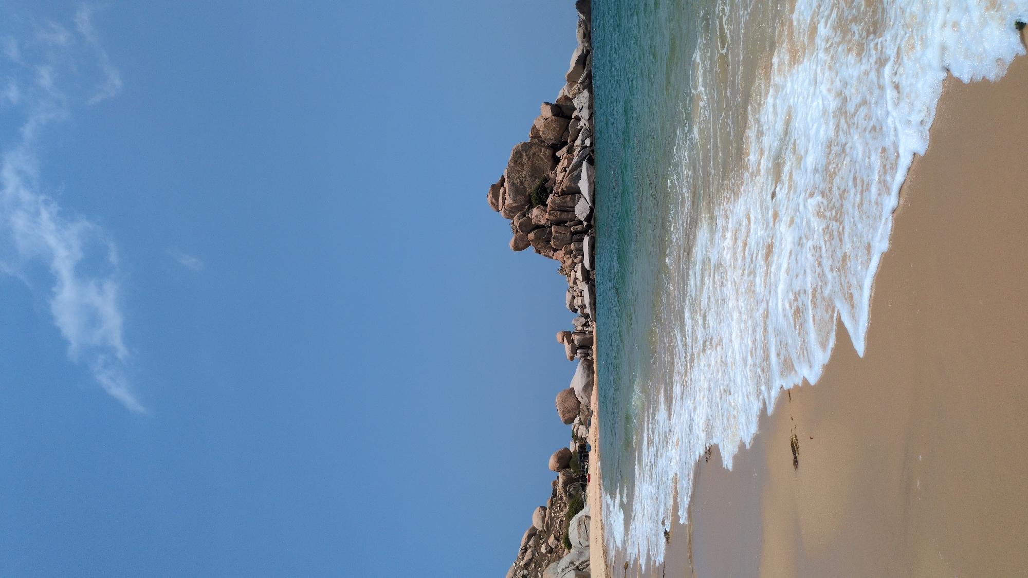 The image is of a beach scene in Phước Dinh, Vietnam. The image is rotated 90 degrees. A clear blue sky with wispy clouds occupies the left side of the frame. The right side depicts a sandy beach with ocean waves washing ashore. A rocky outcrop runs along the edge where the beach meets the water, separating it from the beach. There are people among the rocks and on the beach.