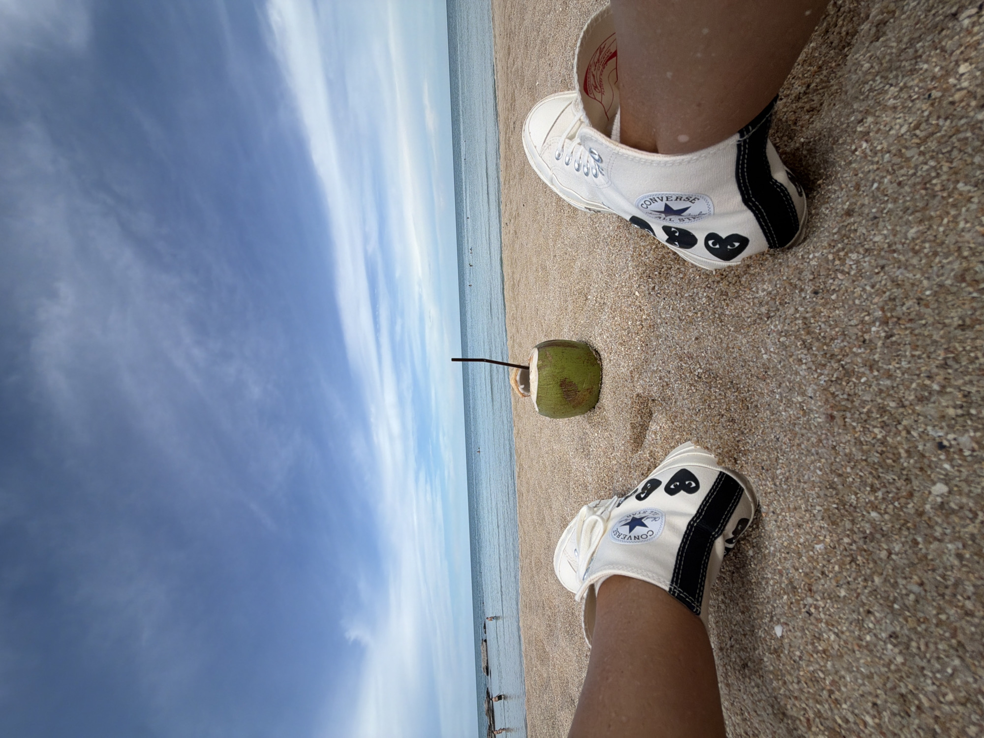 A first-person perspective shot from the beach in Baan Chaweng, Thailand, shows the subject's legs and feet wearing white Converse high-top sneakers with black heart accents. In the background, a green coconut with a straw sits on the sand next to the ocean, with a few people visible wading in the shallow water. The sky above is blue with scattered white clouds, suggesting a pleasant, sunny day. The sand is coarse and tan, typical of a tropical beach.