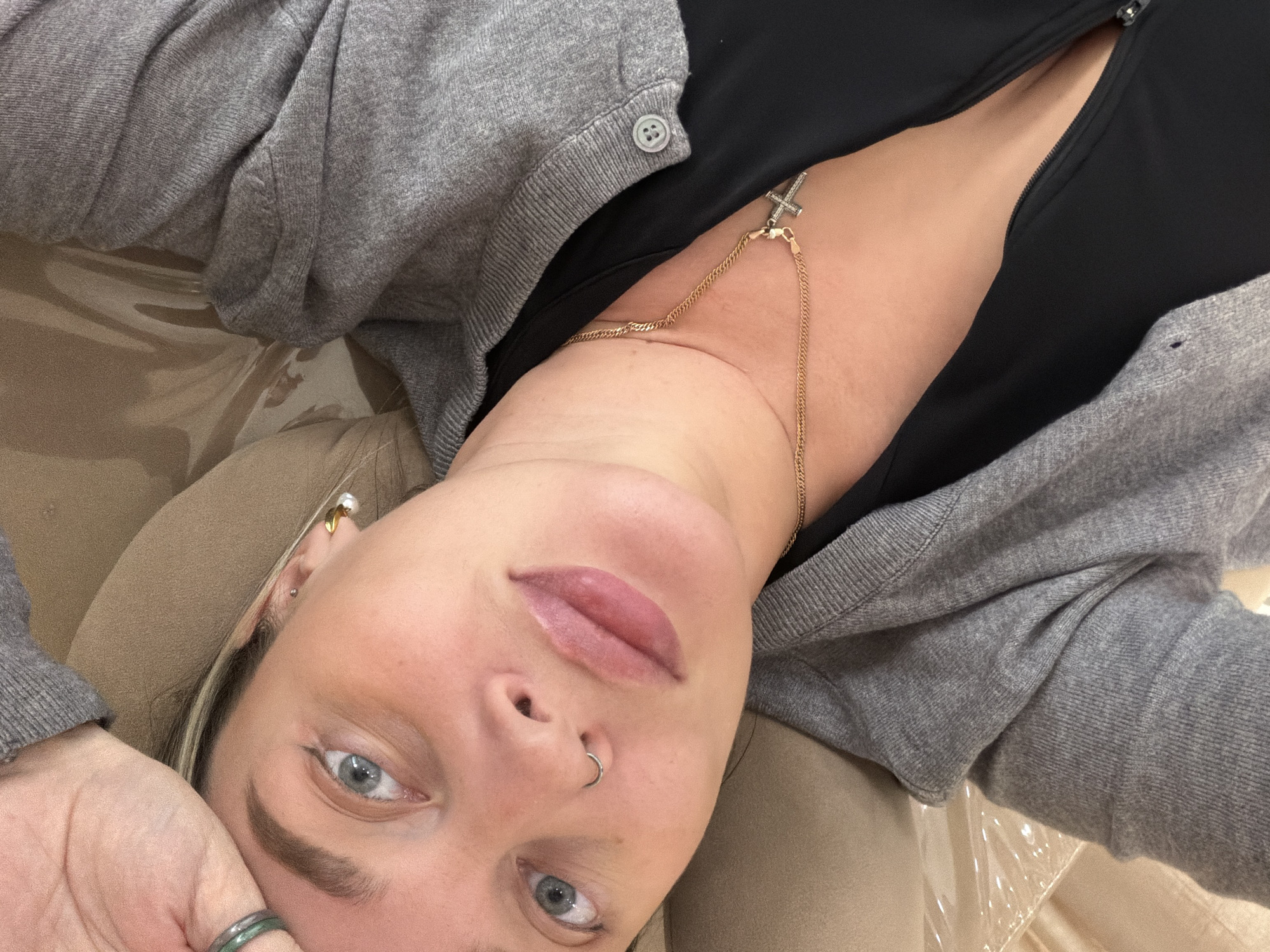 A person is lying down, facing the camera at an angle. The person is wearing a grey cardigan and a black top with a gold chain necklace with a cross pendant. They have blue eyes and a nose piercing. They appear to be indoors. The exact location in Khimki, Russia cannot be determined from the image.