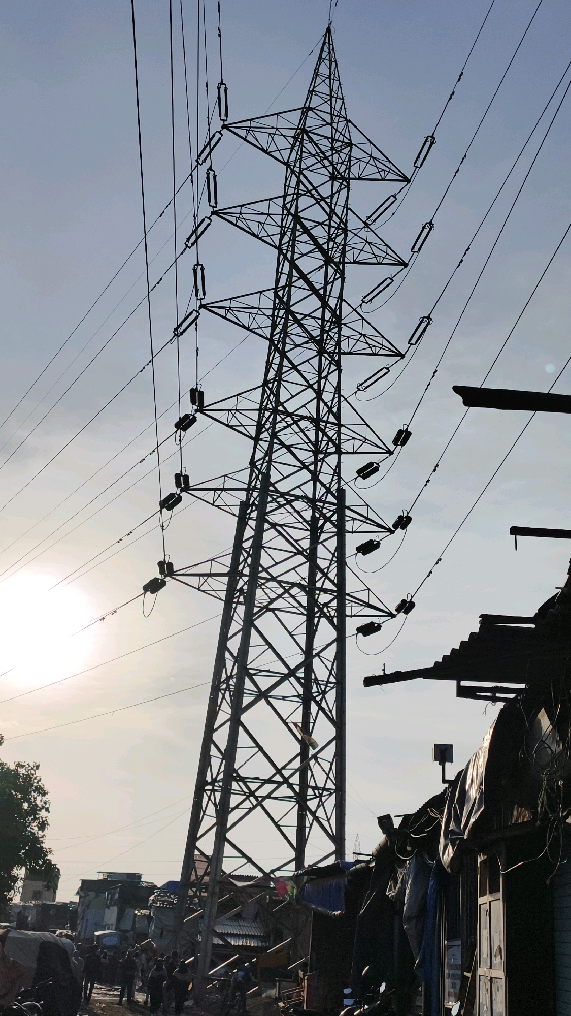 The image depicts a high-voltage electricity pylon dominating the frame against a hazy sky. Several power lines extend from the pylon, suspended by insulators. In the foreground, to the right, is a cluster of makeshift structures with corrugated metal roofs and tarpaulin coverings, suggestive of an informal settlement. To the left and further back, more modest buildings are visible, some appearing to be in disrepair. The bottom of the image shows a group of people gathered, some appearing to be engaged in activities, with motorcycles parked nearby. The presence of the pylon and the dense, informal housing indicates a densely populated urban environment, consistent with areas like Dharavi in Mumbai, India, known for its large-scale informal settlements and industrial activity, often existing in close proximity to essential infrastructure like power transmission towers. The lighting suggests it is either early morning or late afternoon.