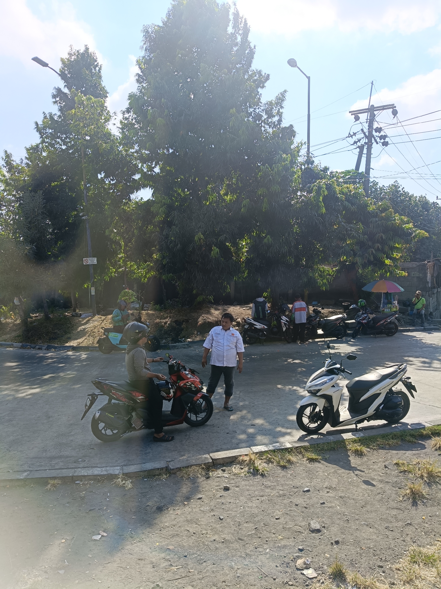 The image depicts a street scene in Kedungrejo, Indonesia, on a sunny day with a clear sky. In the foreground, a person wearing a gray long-sleeved shirt and black pants sits on a red motorcycle. Another person, dressed in a white short-sleeved shirt and dark pants, stands near the motorcycle, looking towards the viewer. To the right, a white scooter is parked on the side of the road.
In the middle ground, several other motorcycles are parked under the shade of large trees. A few people are gathered around the motorcycles, some standing and some appearing to be in conversation. One individual, wearing a white shirt with orange accents, stands near a group of motorcycles. Another person in a white shirt and dark pants is visible further back. A small, colorful umbrella is set up near some of the parked vehicles, suggesting a vendor or a resting spot.
The background features more trees, a utility pole with electrical wires, and a low wall. The ground is a mixture of asphalt and dirt. The overall impression is a moderately busy street or parking area in a tropical or sub-tropical environment.