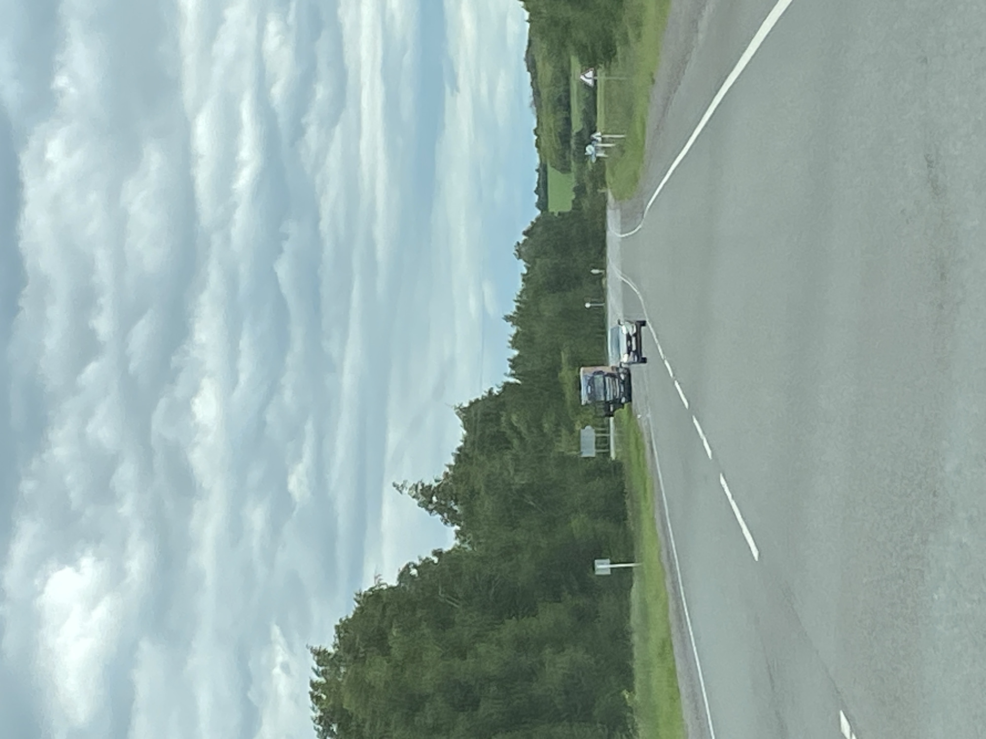 A vertical, eye-level shot captures a two-lane highway in rural Russia, under a cloudy sky. The road stretches into the distance, with white dashed lines marking the lanes. To the left, a dense forest of green trees lines the highway. On the right side, the landscape transitions to rolling green hills with fields in the distance. A silver car is traveling in the left lane ahead, and a large truck is in the right lane further up the road. A few traffic signs are visible on the side of the road. The overall scene suggests a quiet, overcast day in the countryside.