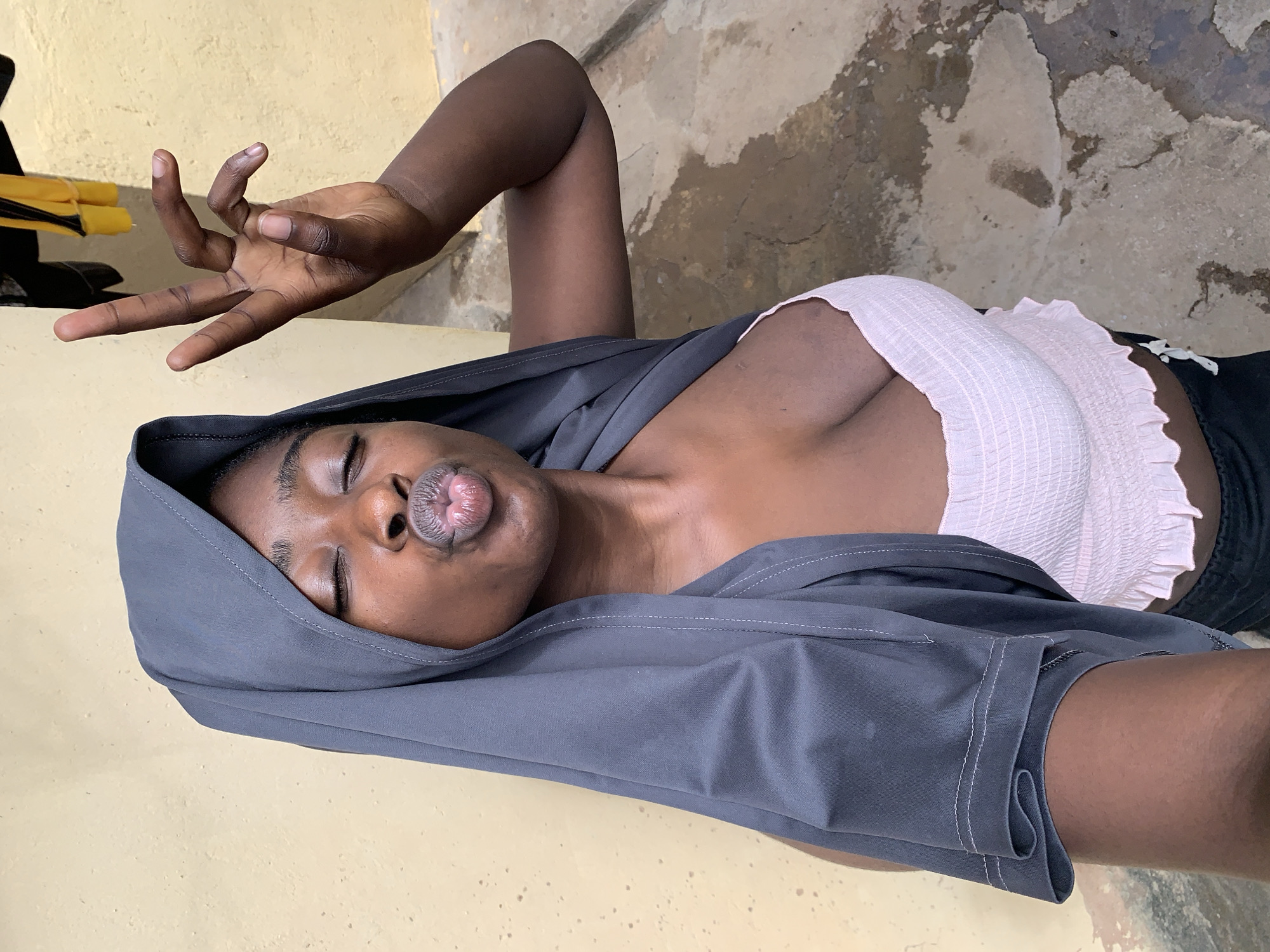 The image shows a Black woman lying down, wearing a light pink top, black shorts, and a dark grey hooded shirt or jacket. Her eyes are closed, and she is puckering her lips. Her right arm is raised with her fingers extended. The background is a beige surface, potentially a wall or floor, and a cracked, mottled area of a different color. The location is identified as Rafin Par, Nigeria. Additional details include yellow objects visible in the background to the left.