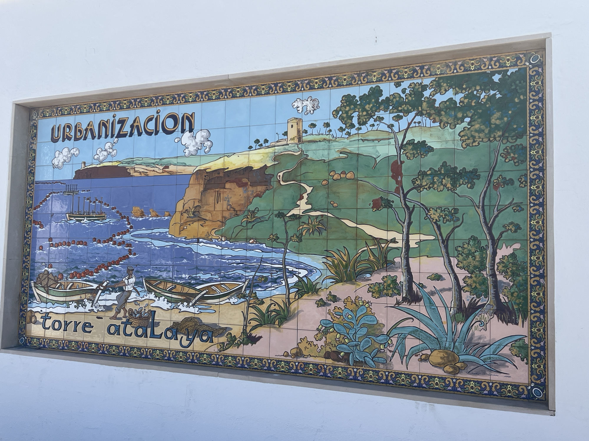 This is a ceramic tile mural depicting a coastal scene with the text "URBANIZACIÓN TORRE ATALAYA". The mural shows a lighthouse or watchtower on a cliff overlooking the sea, with a boat on the water and two smaller boats on the shore. A fisherman is depicted pulling a boat ashore, with a basket of fish nearby. The landscape includes trees, bushes, and rocks. The bottom border of the mural has the inscription "CERAMICAS ARCOS 1992". This mural is located in the Torre Atalaya urbanisation in Conil de la Frontera, Spain.