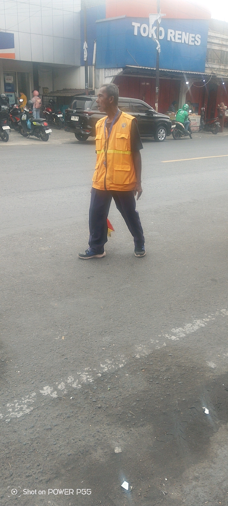 In an outdoor, urban environment, a man wearing a bright orange safety vest over a dark shirt and dark pants walks across an asphalt road. He is captured from the chest down, with his face visible in profile. The man appears to be holding something in his left hand, and a cigarette is visible in his right hand. In the background, a black SUV is parked, and several motorcycles are also present. A blue building with white lettering that reads "TOKO RENES" is visible behind the vehicles. The scene suggests a typical streetscape in a city, possibly during daylight hours, with general activity.