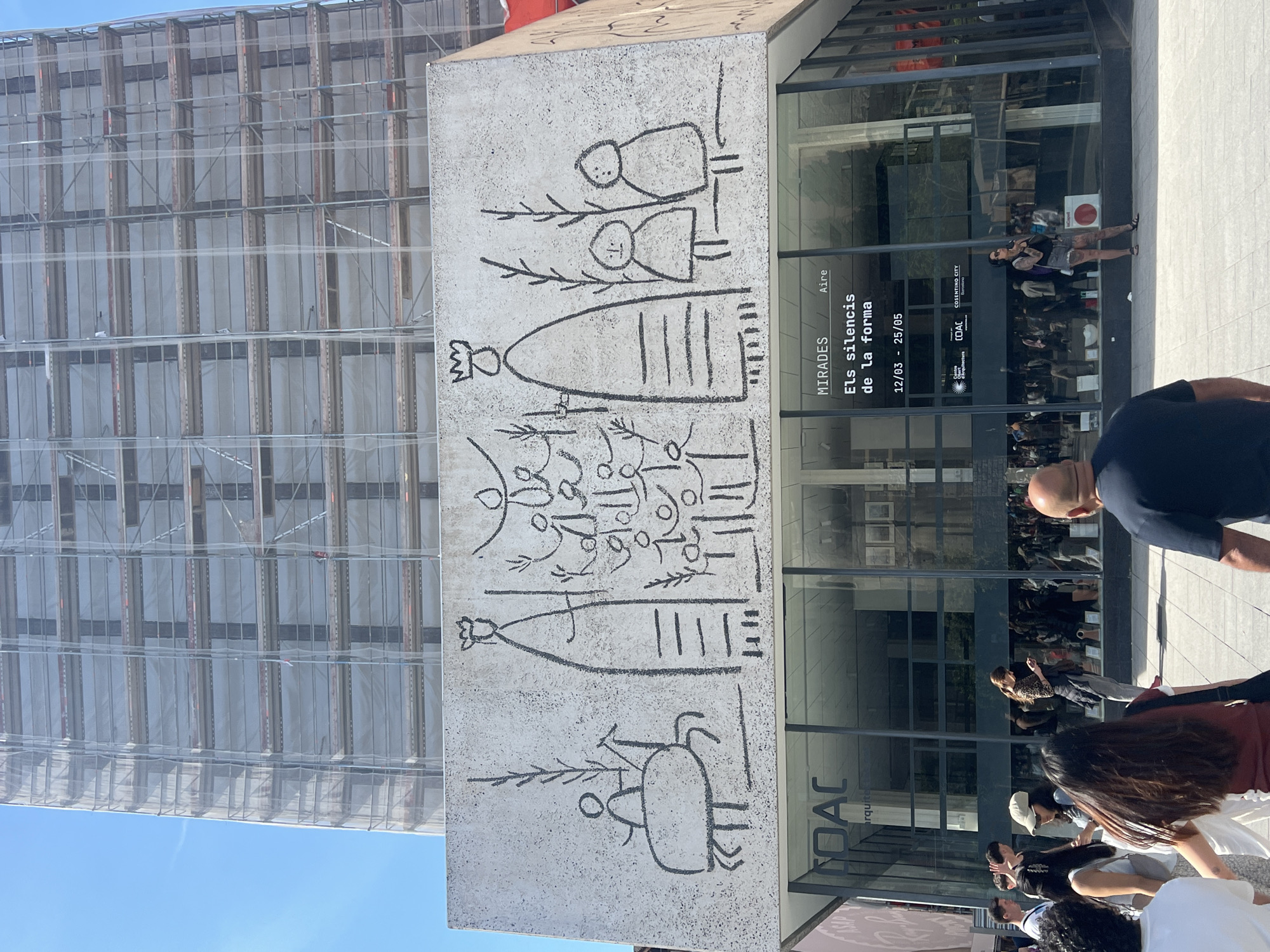 An outdoor scene in an urban environment, likely Barcelona, features a large, textured concrete block in the mid-ground adorned with primitive-style, dark grey artwork. The artwork depicts stylized human-like figures, some with crowns, and abstract organic shapes reminiscent of plants or symbols.
Behind and to the right of this concrete block is a modern building with a reflective glass facade. Through the glass, and in its reflections, several people are visible, some standing, some walking. In the immediate foreground, a man with a bald head, wearing a dark blue shirt, is seen from behind, walking away from the viewer.
Visible text on the glass facade advertises an exhibition: "MIRADES", "Aire", and "Els silencis de la forma", with dates "12/03 - 25/05". Other discernible text includes "COAC", "COSENTINO CITY", and "arquitectura", suggesting the location is likely the Collegi d'Arquitectes de Catalunya.
To the left of the concrete block, a tall building is entirely enveloped in scaffolding and translucent white netting, indicating construction or renovation. A clear blue sky is visible above this structure. The scene is brightly lit by the sun, suggesting midday or early afternoon on a clear day. People appear to be casually moving around, likely passersby enjoying the sunny weather.
