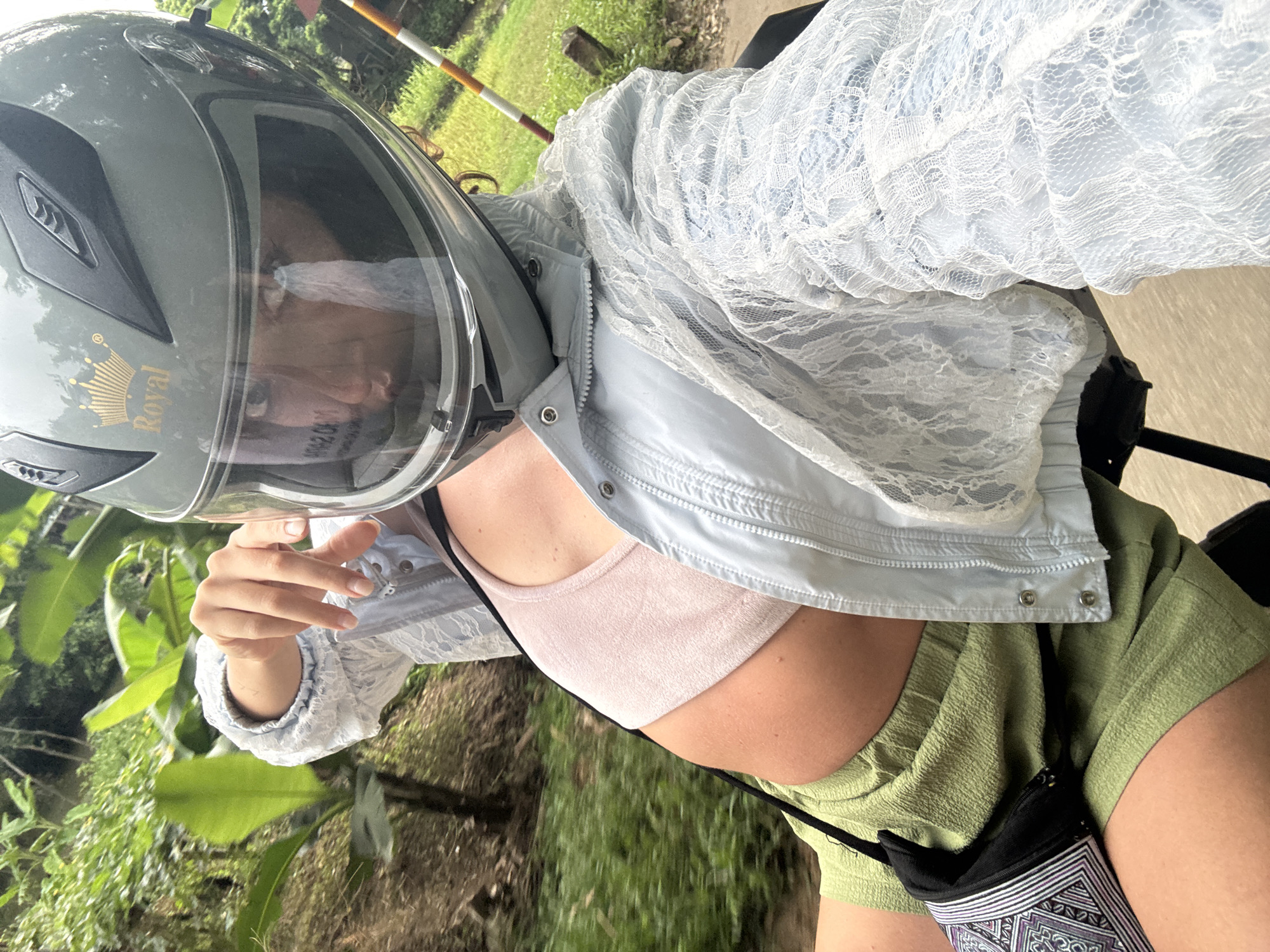A person wearing a light blue lace jacket over a pink crop top and green shorts rides a motorcycle in Nà Mèo, Vietnam. The person has their right hand raised, touching the edge of their grey helmet. The helmet has a gold crown emblem. Green foliage and a rustic fence are visible in the background, along with a blurry road. A small black bag with a patterned strap is slung across the person's body.