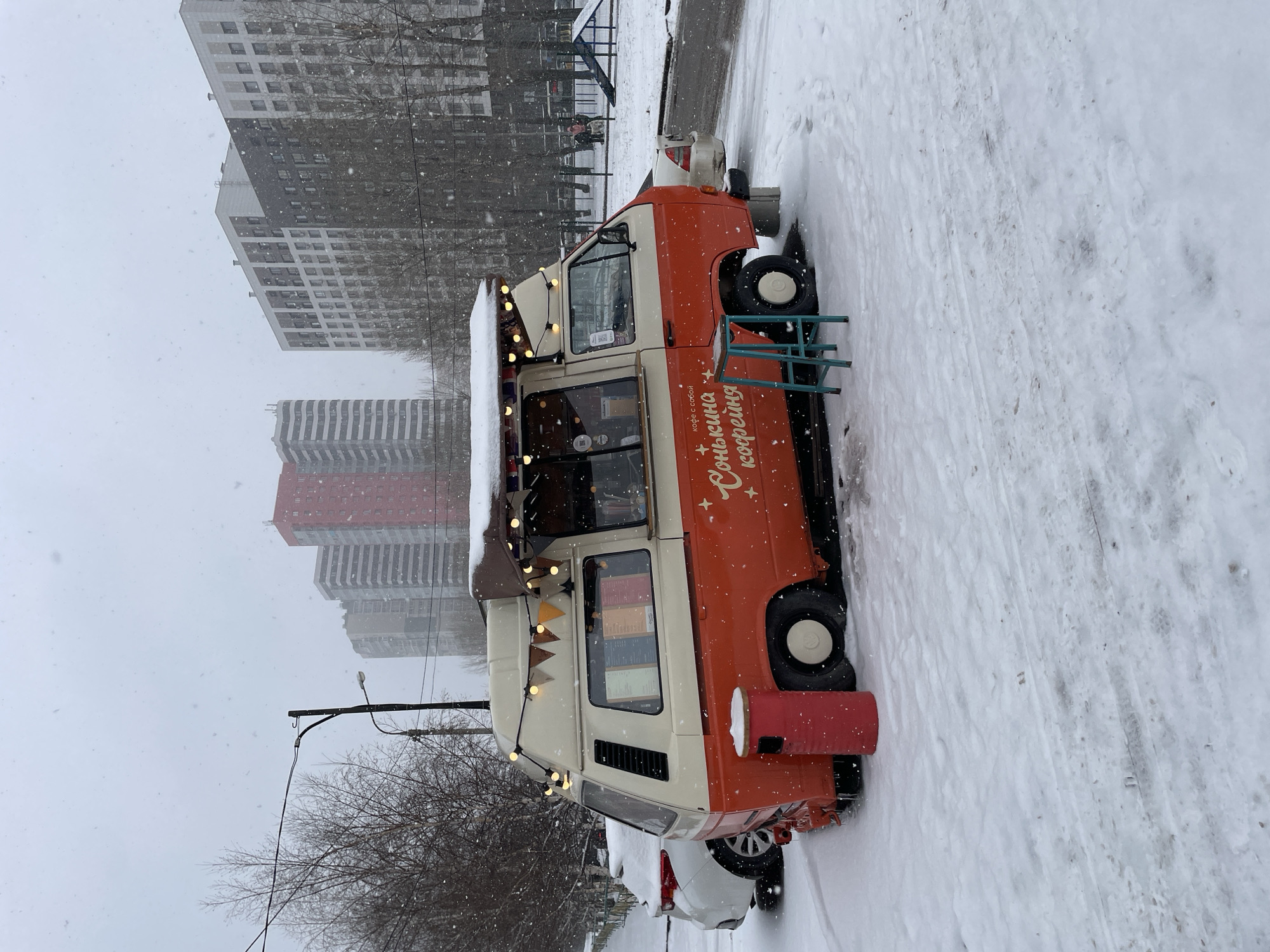 This vertical image captures a winter street scene during a snowfall, centered on a brightly colored food truck. The truck is a vintage-style van, painted cream on top and orange-red on the bottom, adorned with string lights along its awning. On its side, Cyrillic text reads "Солнышко кофейня" (Sunshine coffee shop) with "кофе с собой" (coffee to go) below it. A small green metal stool or table stands next to the truck, and a red cylindrical object, possibly a trash bin or heating unit, is near its front wheel. Snow covers the ground, the truck's roof, and the general surroundings.
Behind the food truck, a white car is partially visible, also covered in snow. The street and sidewalk are a mix of fresh snow and trampled, dirty ice. In the background, towering residential buildings with numerous windows rise against an overcast, grey sky. Some buildings are grey, while one further back shows reddish-brown facades. Bare trees line parts of the street, their branches stark against the sky. Snowflakes are visibly falling across the entire frame, contributing to the wintry atmosphere. No people are visible in the scene. The overall impression is one of a quiet, cold, and snowy day in an urban environment in Krasnoyarsk, Russia.