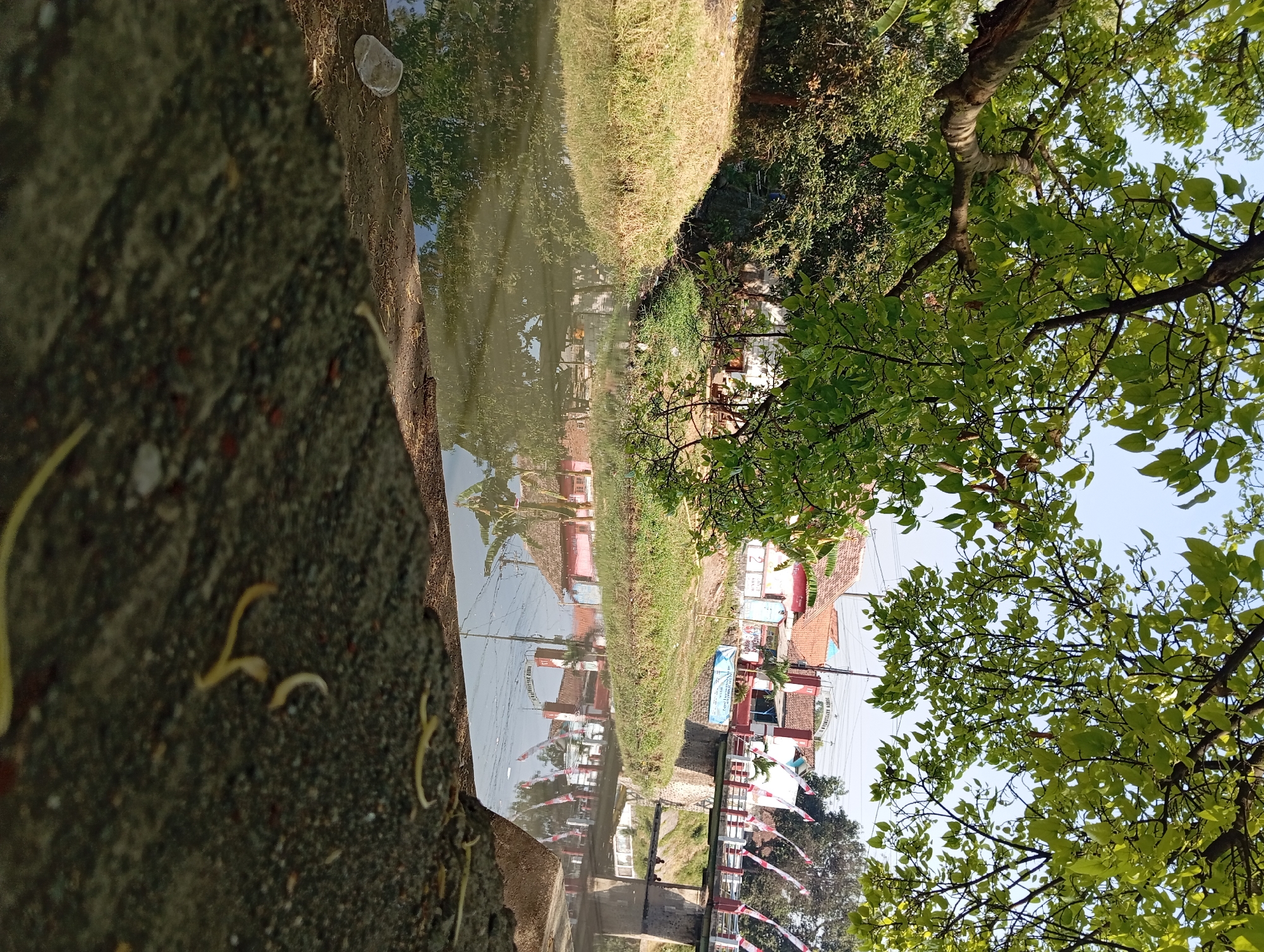 The image depicts a tranquil scene of a river in Kepulungan, Indonesia, under a canopy of lush green leaves. The river's surface reflects the surrounding environment, including a bridge adorned with Indonesian flags and buildings with terracotta roofs. Two individuals are visible near the bridge, seemingly engaged in fishing or relaxing. The foreground is dominated by a textured concrete surface, possibly a pathway or embankment, with fallen leaves scattered across it. The overall atmosphere is serene, capturing a typical day in a quiet Indonesian town.