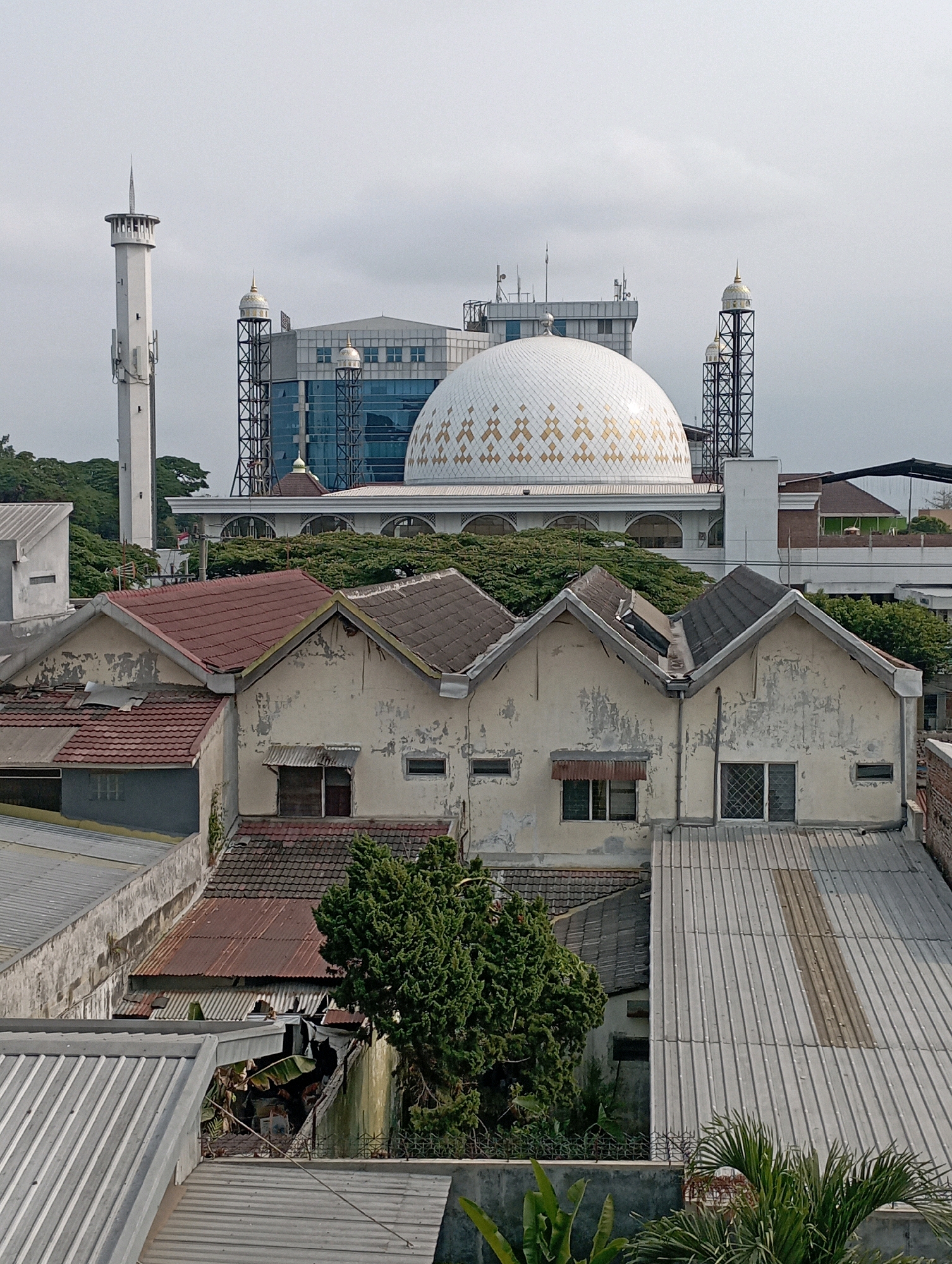 The image shows a view of the Masjid Agung Jami' in Malang, Indonesia. The mosque's prominent white dome with golden geometric patterns is visible, along with two minarets. In the foreground, there are several residential buildings with corrugated metal roofs, some in disrepair, and lush green trees. The sky is overcast. The scene depicts a mix of religious and residential architecture in an urban environment.