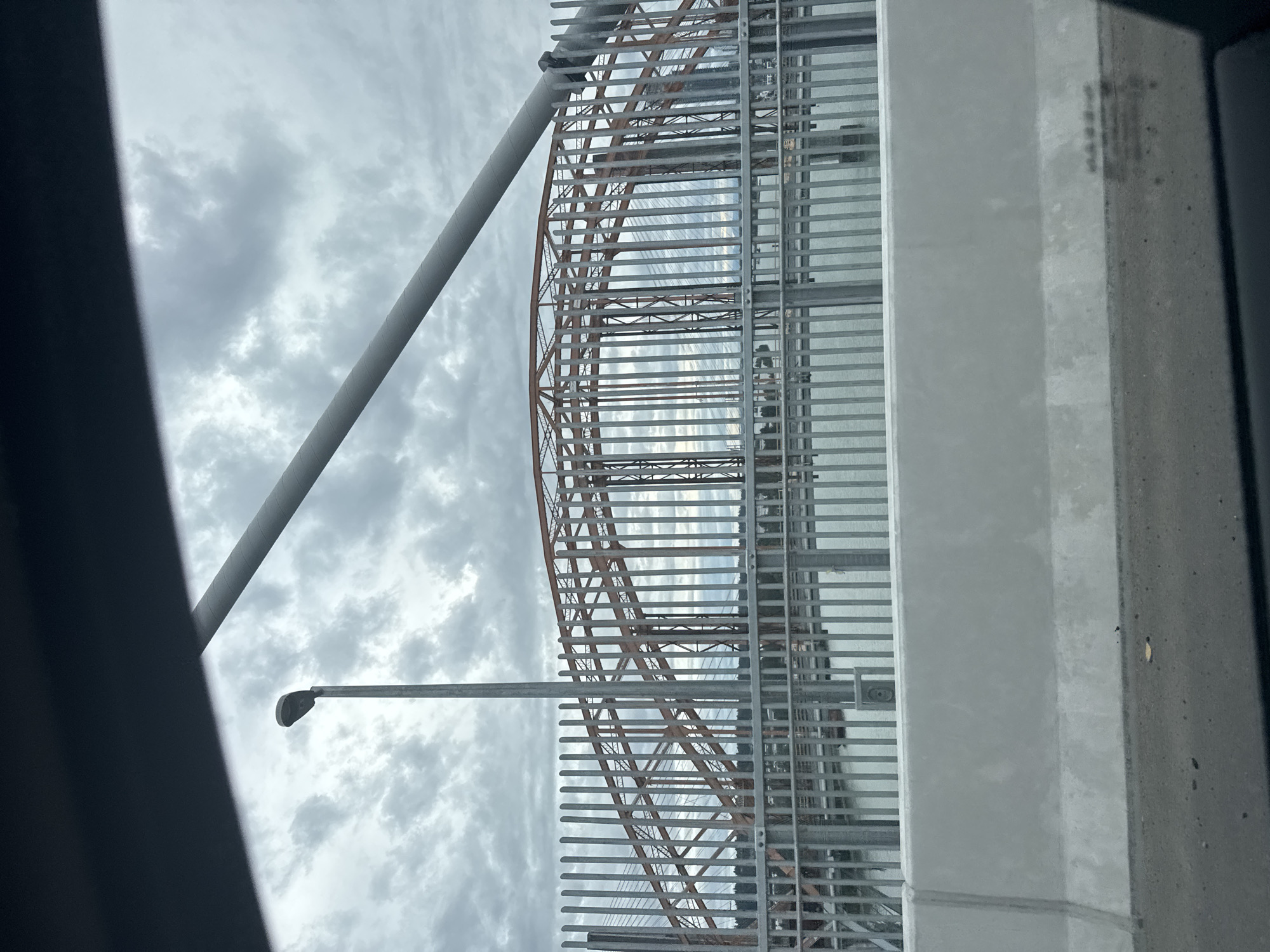 The image shows a view through a vehicle of a bridge under a cloudy sky. The bridge has an orange metal arch structure and is partially obscured by a tall metal fence. A street light is visible to the left of the bridge. The scene appears to be on a bridge or overpass in Surrey, Canada, likely crossing the Fraser River, given the bridge's architecture and its location within Surrey. The foreground shows a concrete barrier and the road surface, indicating the perspective is from a moving vehicle. The water beneath the bridge is calm and reflects the overcast sky. There are no people clearly visible in the image.