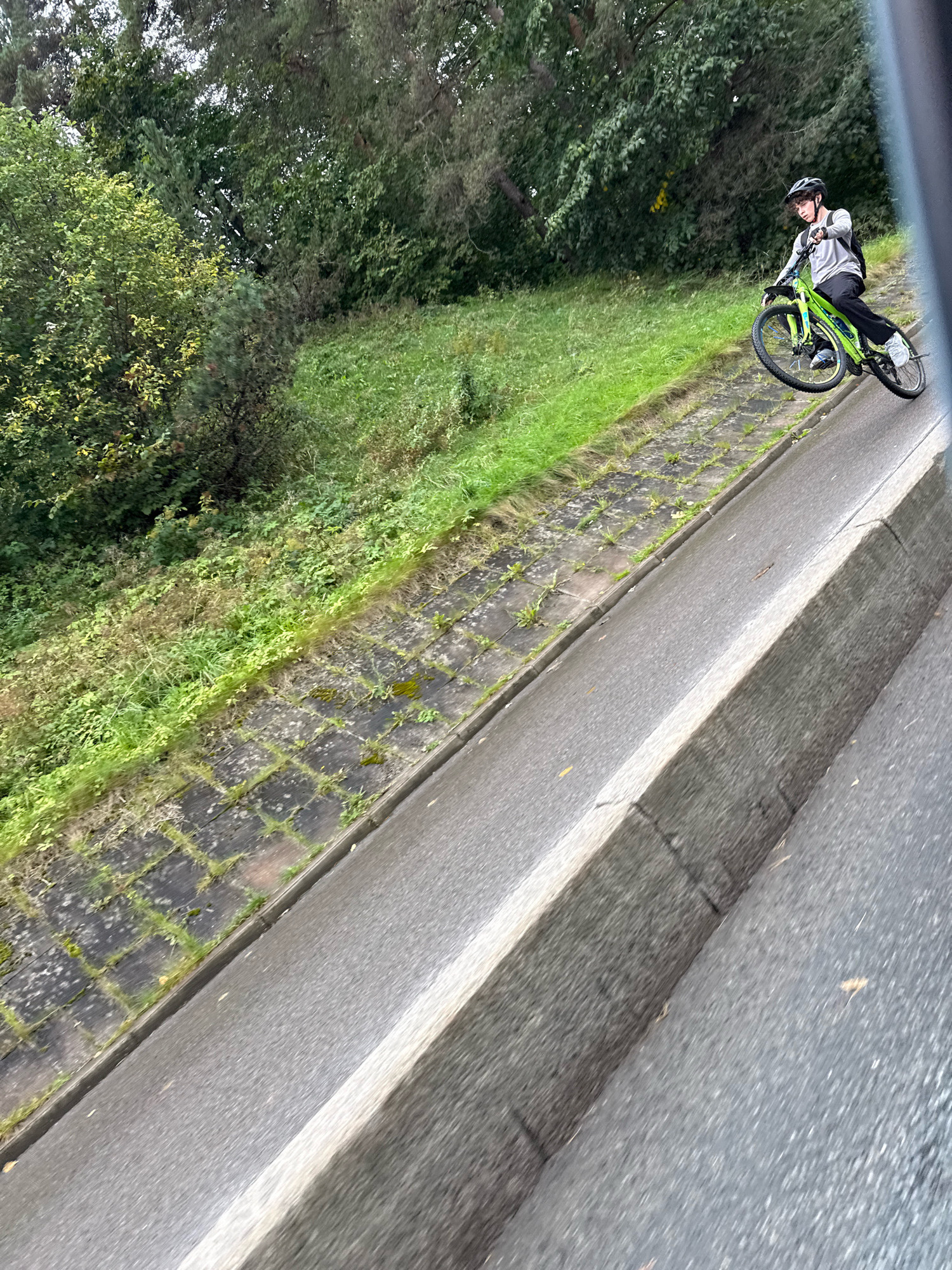 A person wearing a helmet, long-sleeved shirt, and backpack rides a neon green bicycle down a paved embankment. The embankment is lined with stone tiles and overgrown with grass. Trees and foliage are visible in the background. The scene appears to be in an outdoor urban environment, possibly a park or a scenic route.