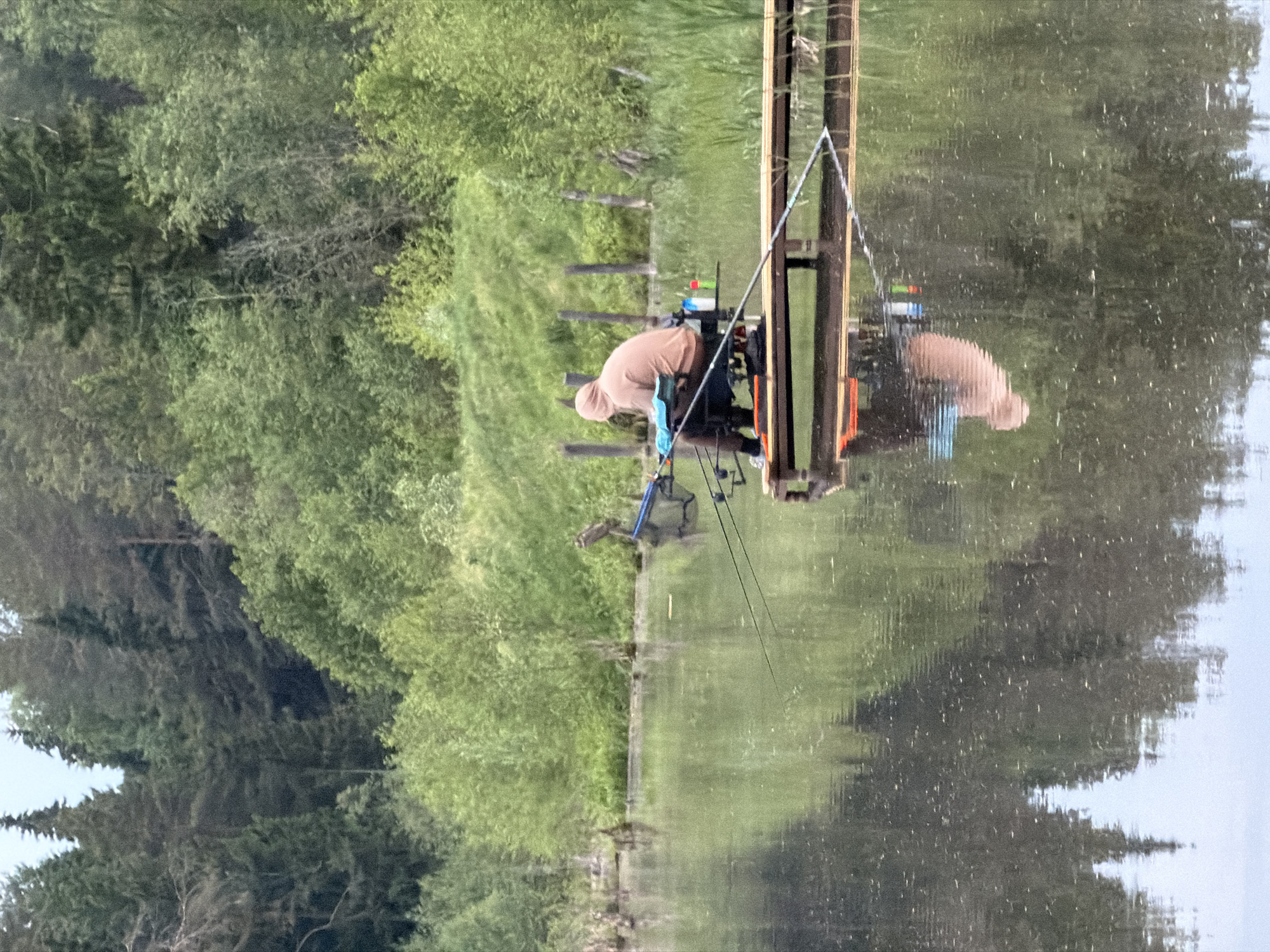 A man in a tan hoodie sits fishing on a wooden platform over the water of a body of water, possibly a lake or river, in Prikamskii, Russia. He is hunched over, possibly attending to his fishing gear. Multiple fishing rods are visible. The water reflects the platform, the man, and the trees on the opposite bank. The banks are grassy with trees lining the shore.