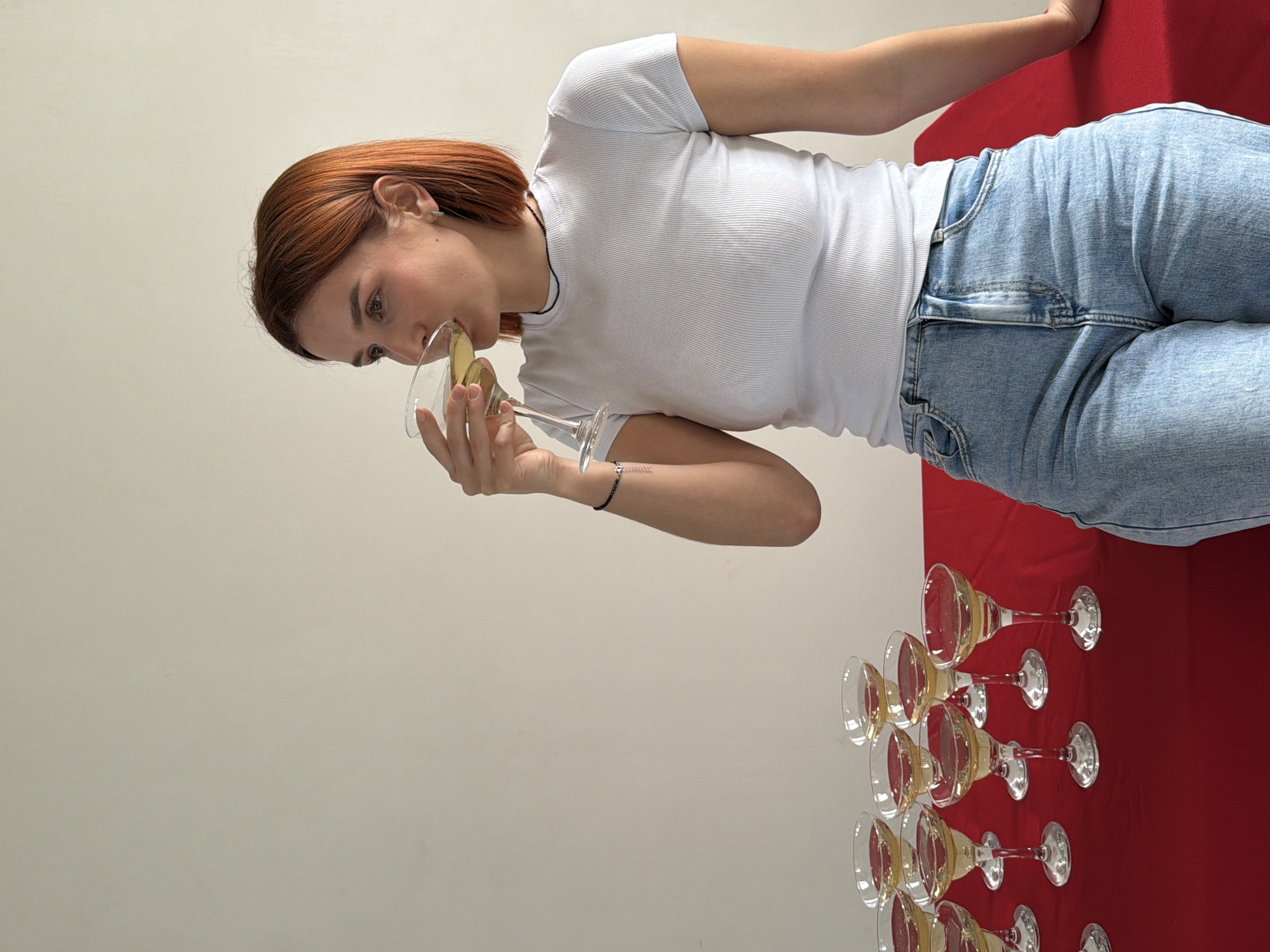 This image captures an indoor scene in Chaikovsky, Russia. A female subject, identifiable by her reddish-brown, shoulder-length hair, light-colored ribbed t-shirt, and blue denim jeans, is positioned towards the right side of the frame. She is depicted holding a martini-style glass filled with a light yellow liquid to her mouth, actively drinking. A thin black necklace and a dark beaded bracelet on her left wrist are visible. Her gaze is directed towards the upper right. To the left of the subject, twelve additional identical martini-style glasses, each containing the same light yellow liquid, are arranged on a red-covered surface. The background consists of a plain, light-colored wall. The subject's right arm rests on the red surface beside her.