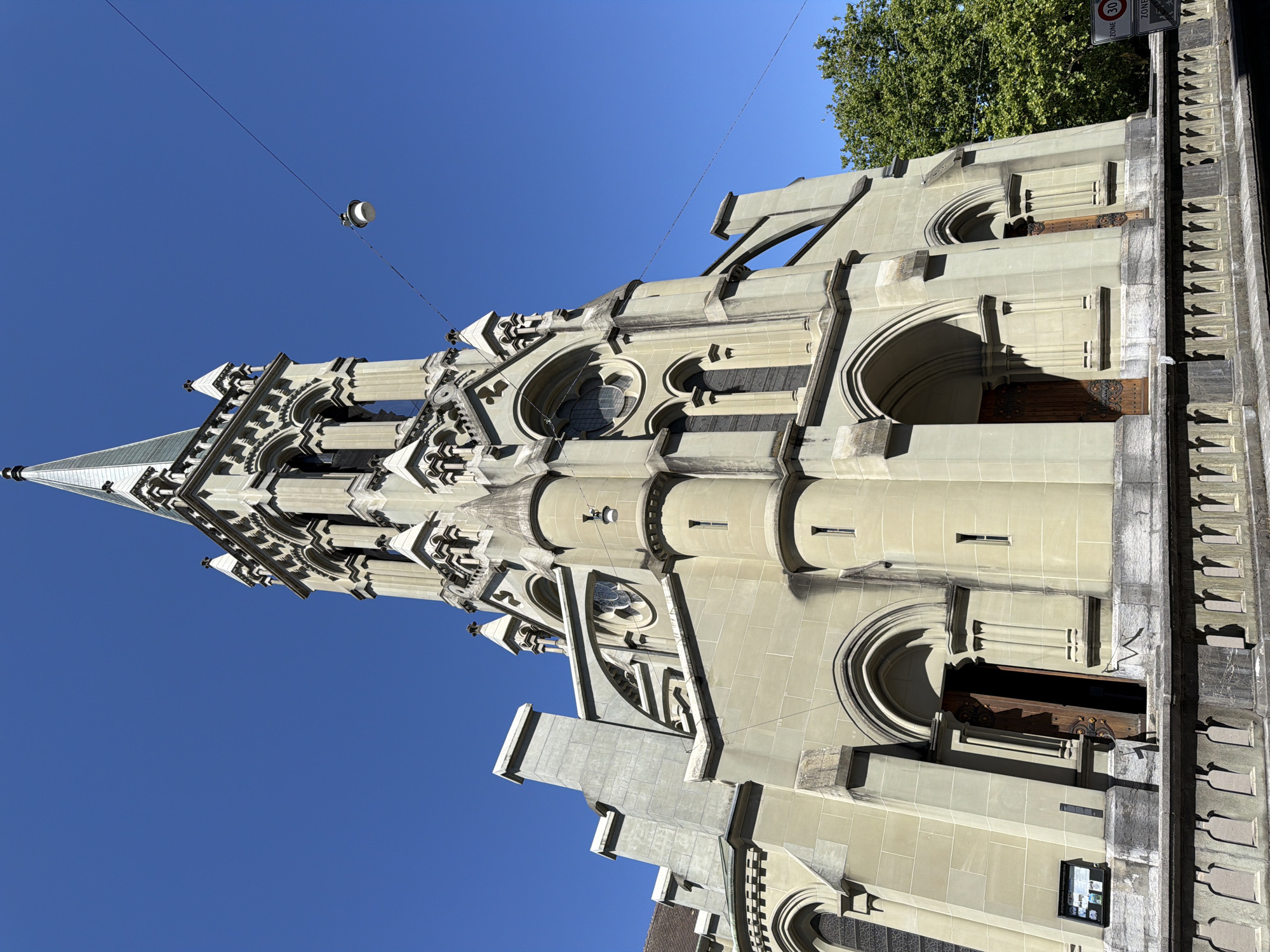The image shows the exterior of the Heiliggeistkirche (Holy Spirit Church) in Bern, Switzerland, on a clear, sunny day. The church is made of light-colored stone and features a prominent bell tower with a spire. The facade has several arched doorways and windows, as well as intricate architectural details. A lamppost hangs from a wire across the sky in front of the tower. In the background, lush green trees are visible, and a street sign indicating a 30 km/h speed limit is partially visible on the right side of the frame. The church appears to be situated on a slightly elevated position, with a stone balustrade in the foreground. There are no people visible in the image.