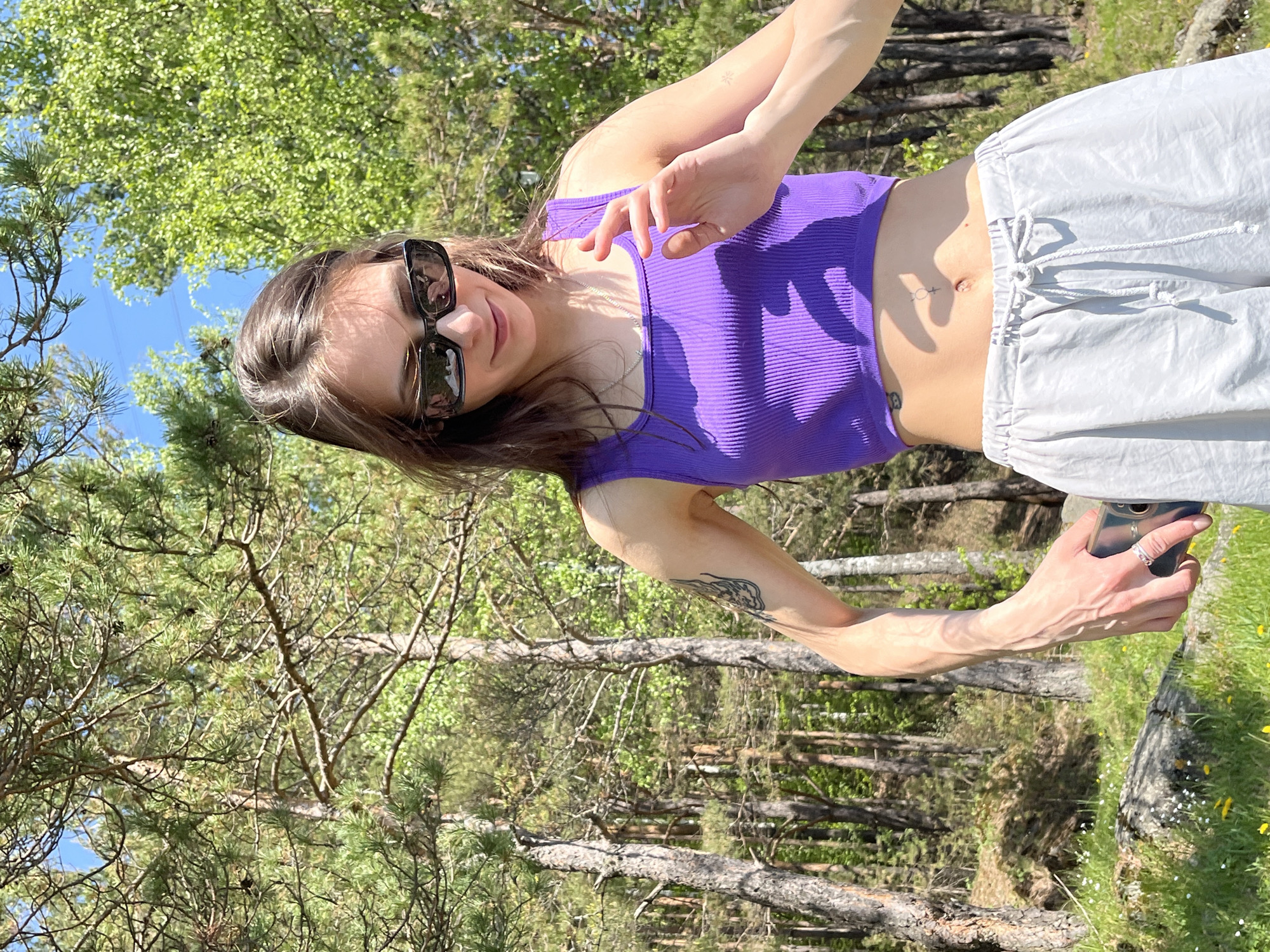 A young woman is captured in an outdoor setting in Слизнево, Russia, appearing to take a selfie or record a video with her smartphone. She is positioned slightly angled, with her head and torso rotated towards the camera, and her right arm extended forward holding the phone. She wears dark rectangular sunglasses, a ribbed purple crop top, and light grey or white drawstring pants. A silver chain necklace is visible around her neck. A small tattoo in the shape of the Venus symbol with an additional horizontal line is visible near her navel, and another tattoo, possibly depicting wings, is on her left forearm. A silver ring adorns her right ring finger.
The background is a vibrant forest or wooded area, dominated by tall pine trees and some deciduous trees with lush green foliage, suggesting spring or summer. The sky above is clear and blue. Bright sunlight casts distinct shadows, indicating a sunny day, likely during the mid-day hours. The ground is covered with green grass, and small yellow and white wildflowers are visible in the foreground to the right. Several fallen tree trunks and branches are scattered throughout the scene. The woman has a slight, relaxed smile. No discernible text is visible on her clothing or the immediate surroundings.