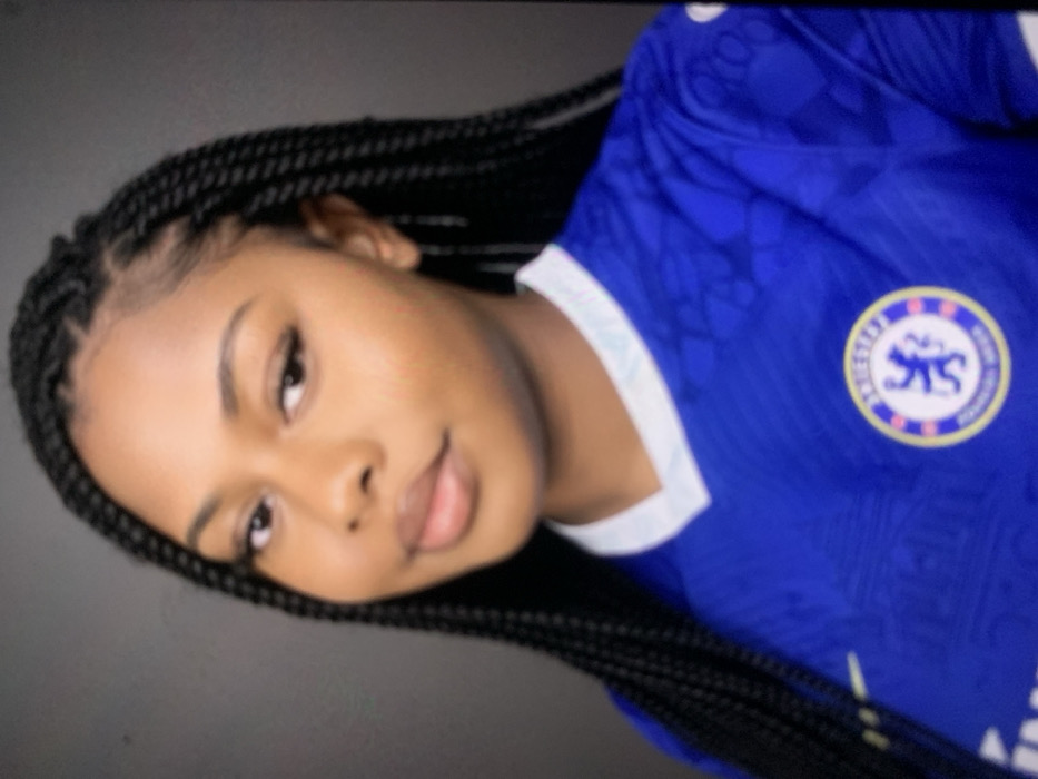 A medium shot captures a young adult female with dark skin, facing slightly towards the camera with her head tilted to the viewer's right. Her hair is styled in numerous small, dark box braids that extend down past her shoulders. She is wearing a royal blue Chelsea Football Club jersey, featuring the club's crest with a lion rampant on the viewer's left side of the chest, and a white Nike swoosh logo on the viewer's right. The jersey has a white V-neck collar with subtle light blue detailing. Her facial features include full lips and dark eyes, with visible eyeliner. The background is a plain, dark grey, devoid of any specific environmental details. No visual information within the image definitively identifies a location in the city of Jos, Nigeria.