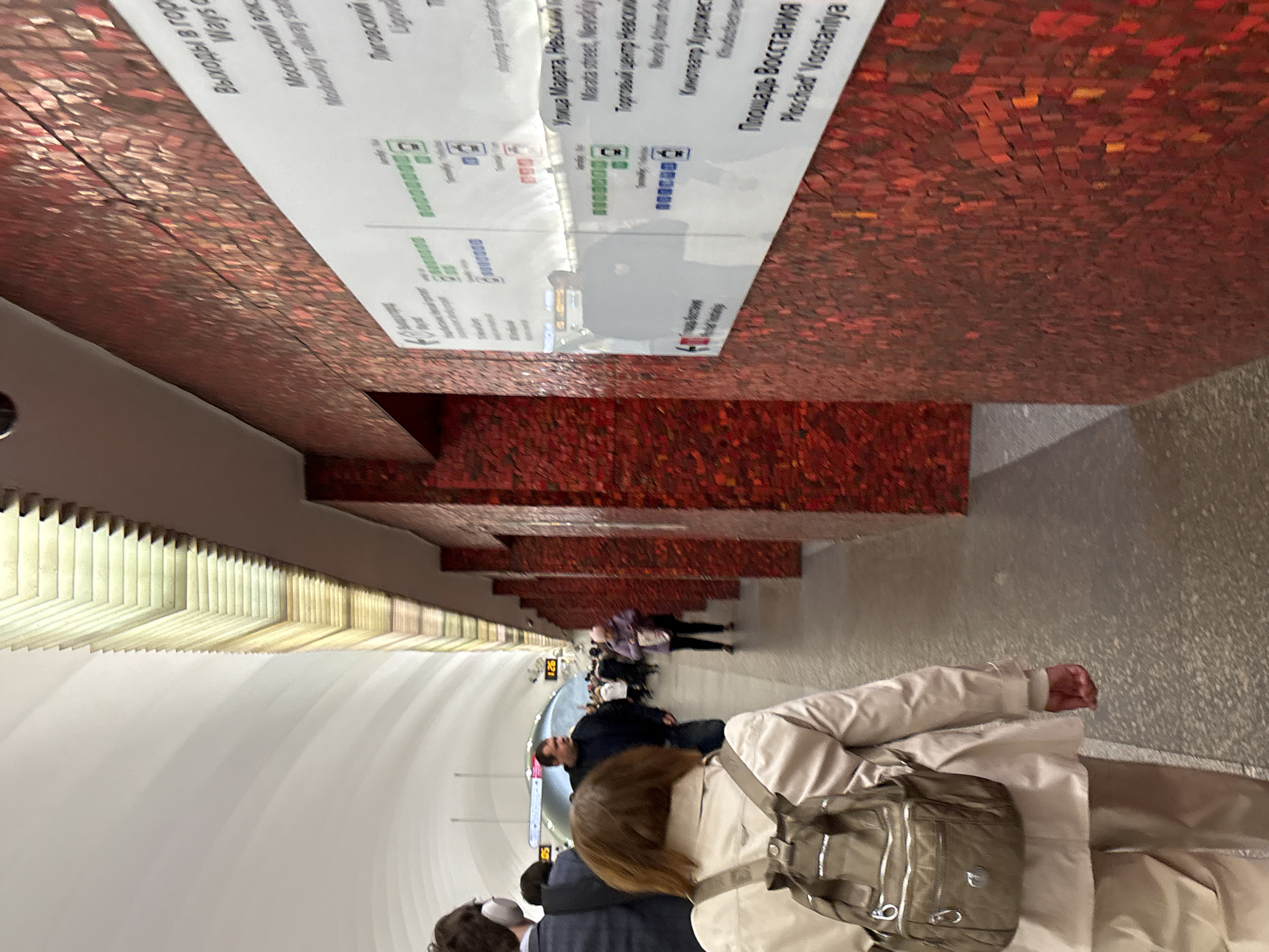 This image captures a scene from a subway station, specifically the "Ploschad Vosstaniya" station in Saint Petersburg, Russia. People are seen walking within the station. The station's interior features a distinctive red-tiled wall design, complemented by illuminated ceiling fixtures. A sign is visible, displaying information about various exits and points of interest like "Moskovskiy railway station," "Ligovsky," "Marata Street, Nevskiy," and "Nevskiy Shopping Center."