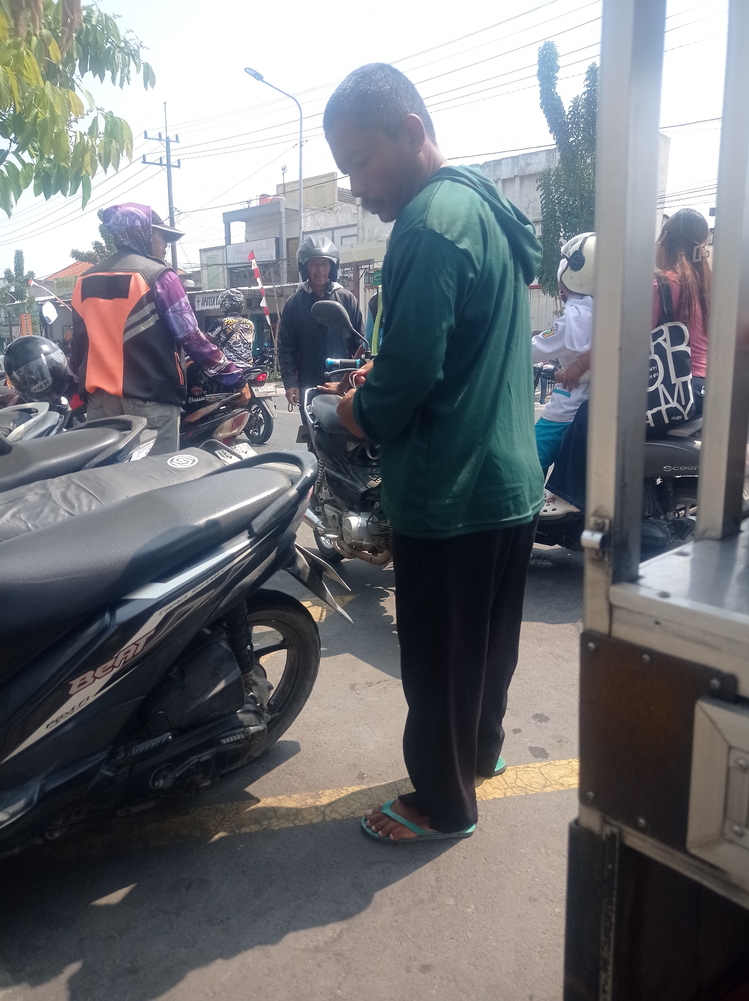 The image depicts a street scene in Bangunsari, Indonesia, on a bright, sunny day. In the foreground, the side of a black scooter, identified by the "BEAT" branding, is visible. To the right, a metal enclosure with rivets and a textured surface suggests a vehicle or structure. A man wearing a green hooded sweatshirt and dark pants stands in the middle ground, facing away from the camera and holding an object. He is wearing turquoise flip-flops. Behind him, several other people and motorcycles are present. One individual on a motorcycle wears an orange and purple vest, a patterned headscarf, and a helmet. Another person on a motorcycle wears a black helmet. Further back, a woman with long brown hair tied back with a clip is visible, wearing a pink shirt and a black backpack with white lettering. The environment includes buildings, utility poles, and trees, indicating an urban or semi-urban setting. The overall impression is that of a busy street or marketplace.