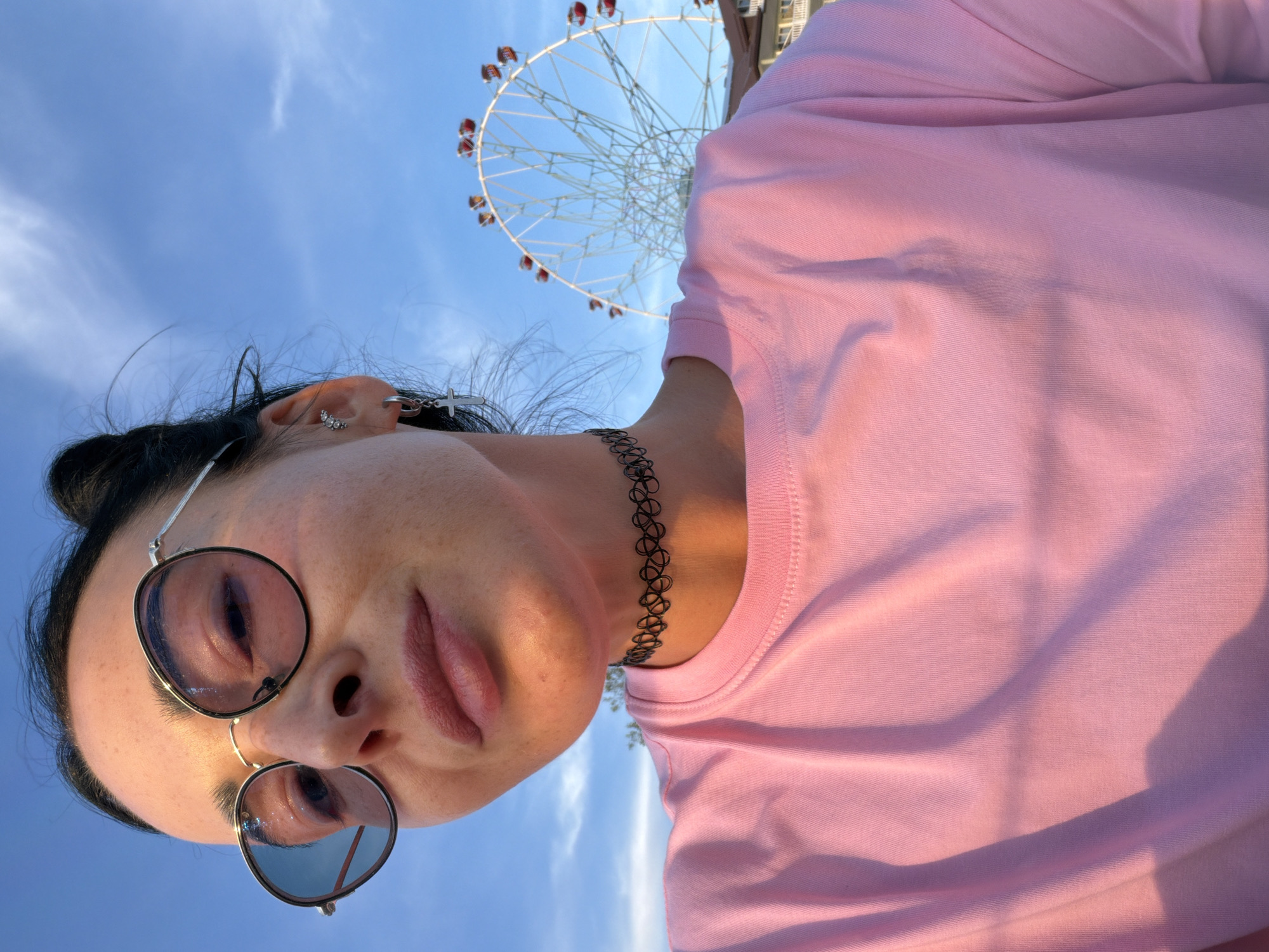 A selfie shows a young woman wearing round, wire-rimmed sunglasses and a black choker necklace. She has dark hair pulled back and is wearing a pink t-shirt. In the background, a Ferris wheel is visible against a bright blue sky. The lighting suggests it is late afternoon or early evening. The Ferris wheel and the architectural style of a building visible behind it are consistent with locations in Sochi, Russia, an area known for its amusement parks and resorts, particularly along the coast. The scene appears to be taken outdoors, possibly at an amusement park or recreational area near the Black Sea coast.