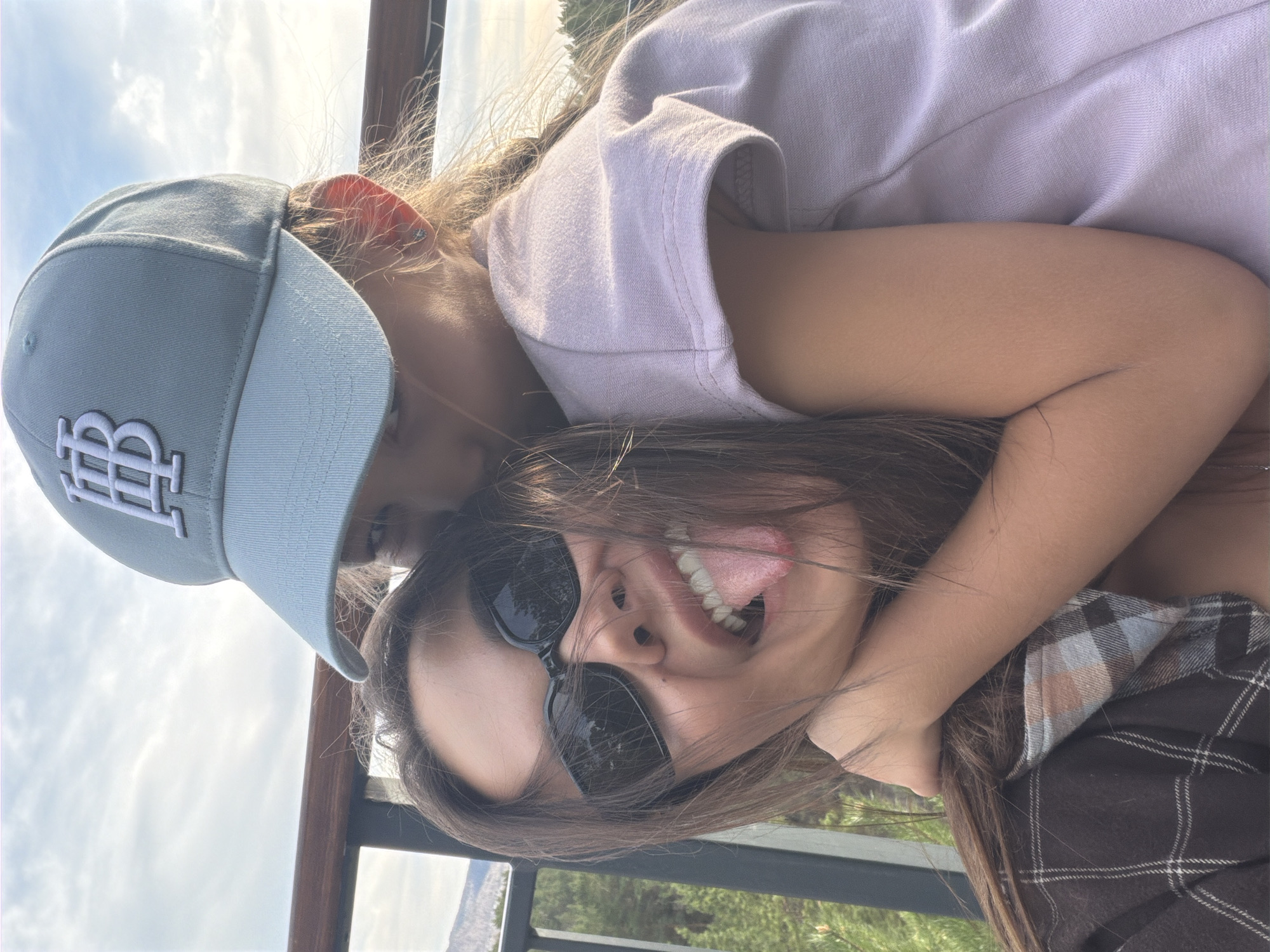 Two young women are in a friendly embrace, one wearing a gray baseball cap with a white logo, a lavender-colored shirt, and has light brown hair. The other, in the foreground, is wearing sunglasses, has dark hair, and is sticking her tongue out while smiling. They appear to be standing outdoors on what may be a balcony or overlook with a metal railing. The background suggests a natural landscape, possibly with trees or hills. The sky is visible and cloudy. The location is in the city of Krasnoyarsk, Russia.