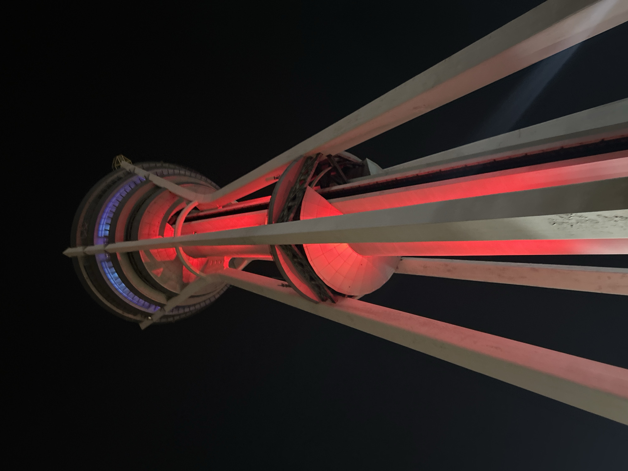 The image shows a low-angle, upward view of the Alor Setar Tower (Menara Alor Setar) at night in Alor Setar, Malaysia. The tower is illuminated with vibrant red lights that shine up its central structure and along its exterior supports. The top section of the tower features blue and purple lighting. The sky is dark, indicating nighttime. The tower's concrete legs are visible extending upwards, supporting a multi-tiered structure at the top. The overall impression is one of architectural grandeur and modern illumination against the night sky.