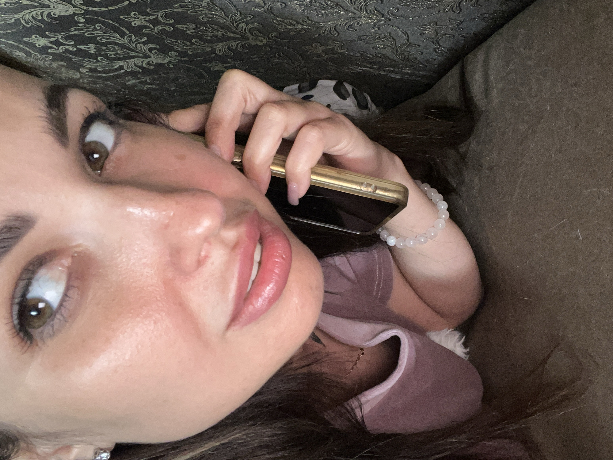 A close-up shot focuses on the face and upper body of a woman who is holding a gold-colored smartphone to her right ear. The woman is looking upwards and to her left with her mouth slightly open. Her skin appears flushed, with visible pores and some sheen. She has brown eyes, dark eyebrows, and long dark hair that is partially visible around her face. She is wearing a pink t-shirt and a beaded bracelet on her left wrist. A patterned fabric, possibly a scarf or clothing, is visible behind her right ear. The background consists of a dark, patterned wallpaper on the right and a textured, dark brown surface that appears to be a cushion or sofa on the bottom. The lighting suggests an indoor setting, possibly at night, with a warm light source illuminating her face from the front.