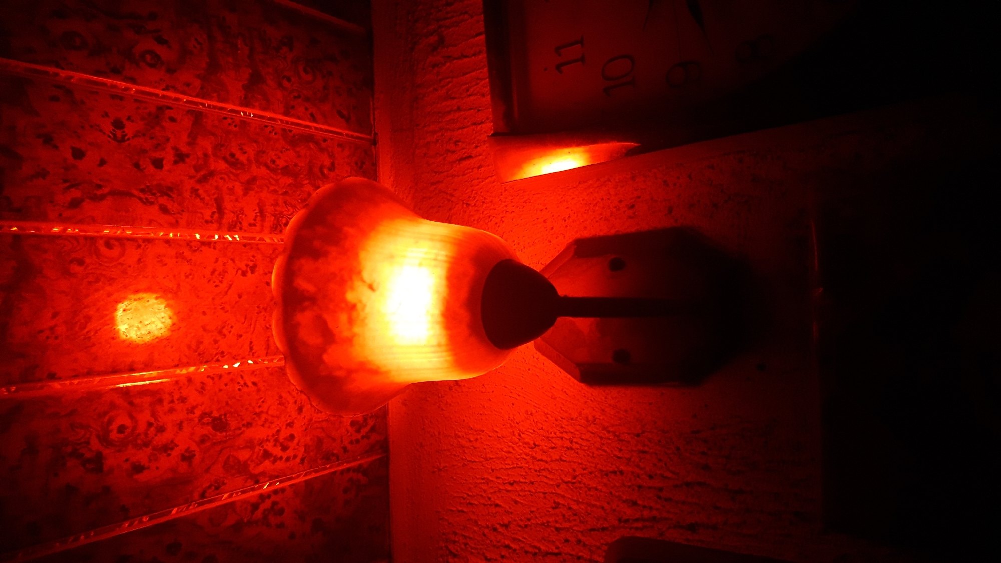 The image depicts a dimly lit interior. A wall sconce with a frosted glass shade glows brightly, casting a red hue throughout the scene. The wall it is mounted on has a textured surface. Above the sconce, a portion of a clock face with the numbers "10" and "11" visible suggests time. To the left of the sconce, a textured or patterned surface appears, possibly a wall covering or decorative panel, with linear elements resembling shelves or moldings. The overall scene is dark, with significant areas of shadow, emphasizing the red glow from the sconce. The location is unknown; Nkpor, Nigeria, cannot be confirmed.