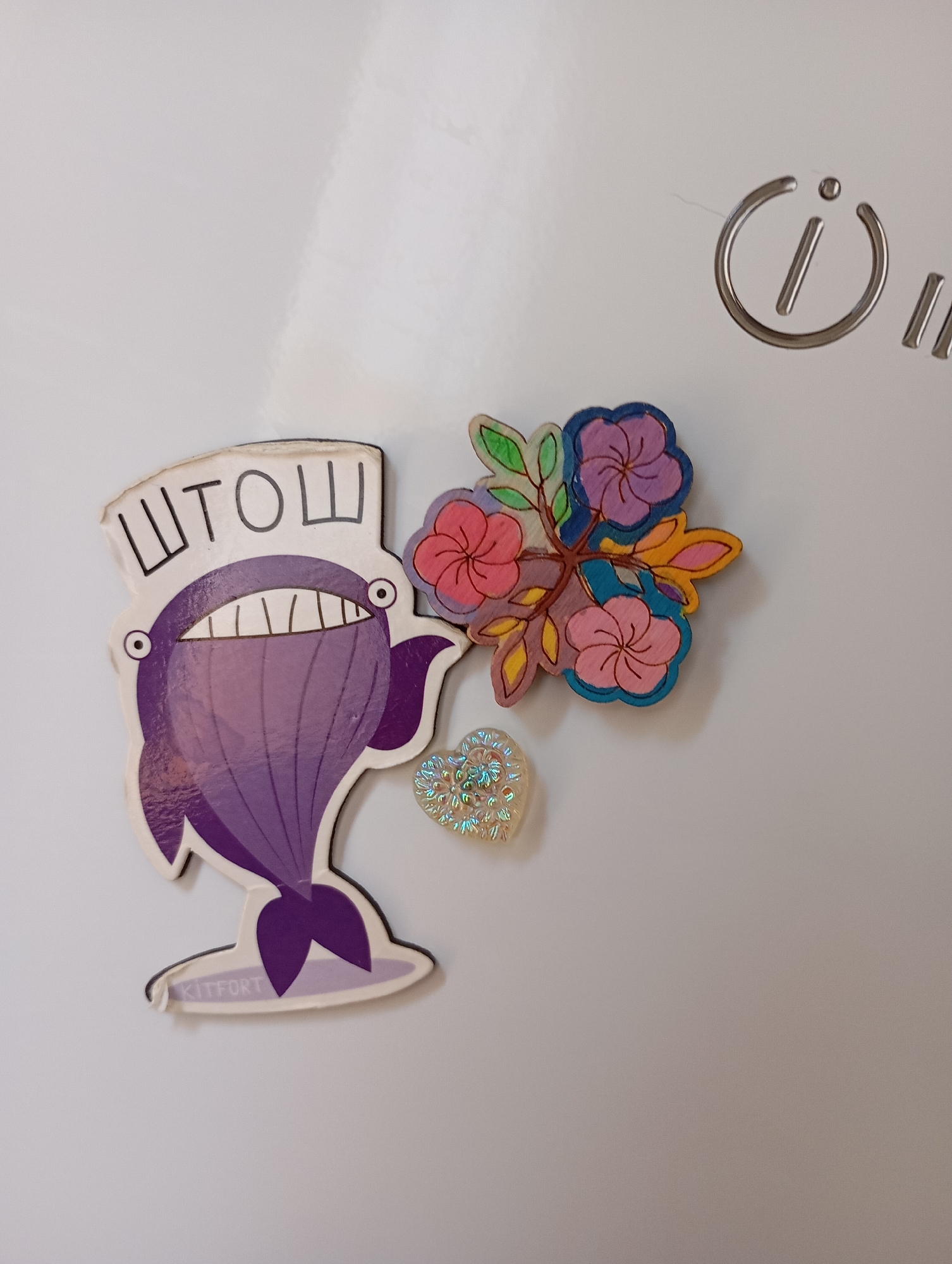 The image shows a white refrigerator door with several magnets. On the left is a purple whale magnet with the Cyrillic letters "ШТОШ" above its head and the word "KITFORT" on its tail. To the right of the whale is a magnet depicting a cluster of colorful flowers, with pink, purple, and yellow petals and green leaves. Below the flowers and to the right of the whale is a small, iridescent heart-shaped charm. In the upper right corner of the refrigerator, a silver power button symbol is visible. The background is a plain white surface, consistent with a refrigerator door. No people or actions are depicted in the image. The location of Gorki-2, Russia, is not visually represented in the image itself.