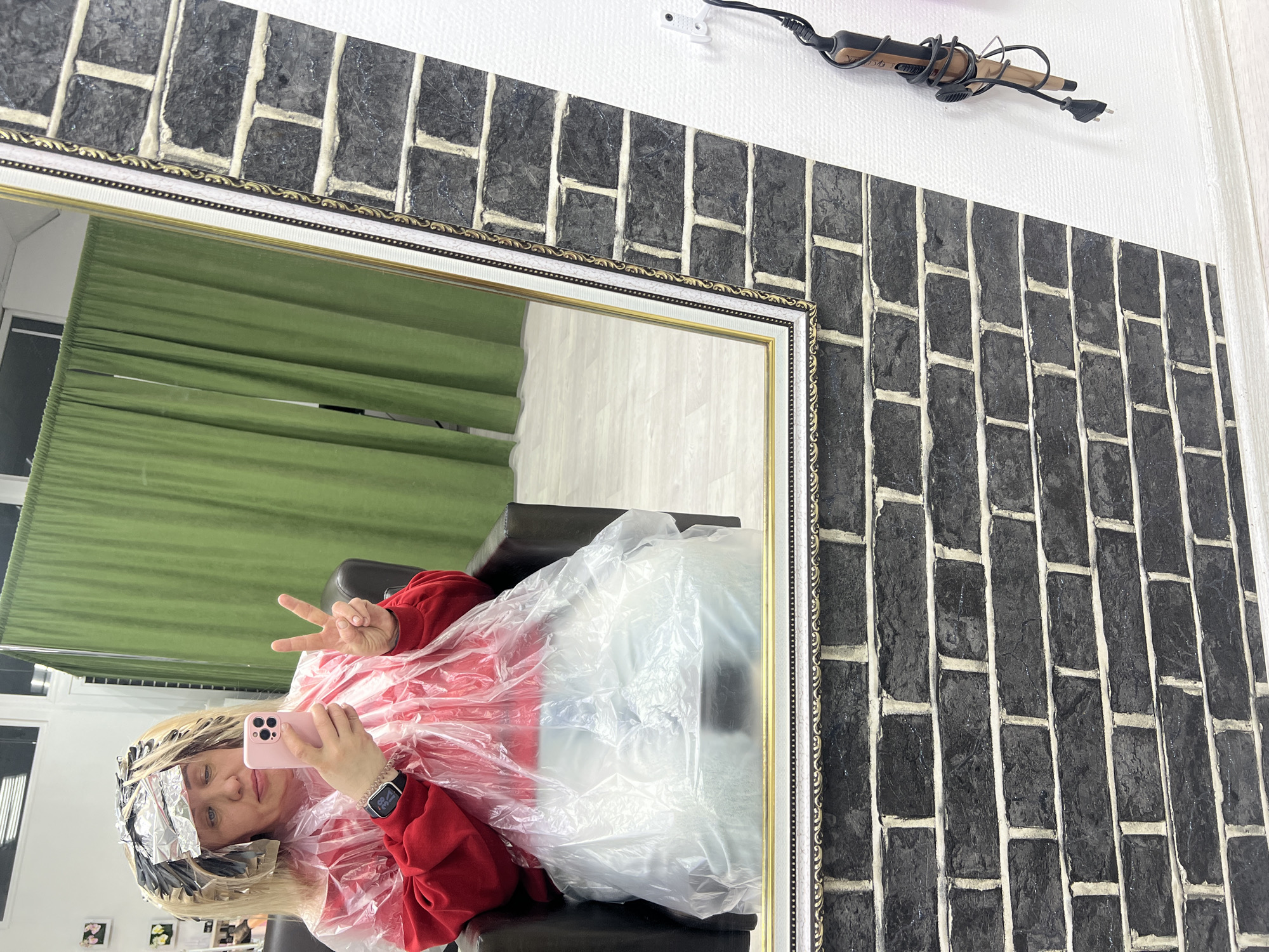 A medium indoor shot depicts a person reflected in a mirror. The individual is seated, wearing a red sweater and a clear plastic cape, with hair partially covered in foil. The person holds a pink cell phone and gestures a peace sign. The reflection suggests the environment includes a green curtain and white walls. The mirror is framed in gold and white. The wall around the mirror is decorated with a black and white brick pattern, and a curling iron is mounted on the upper part of the wall. The location is potentially a salon in Chaikovsky, Russia.