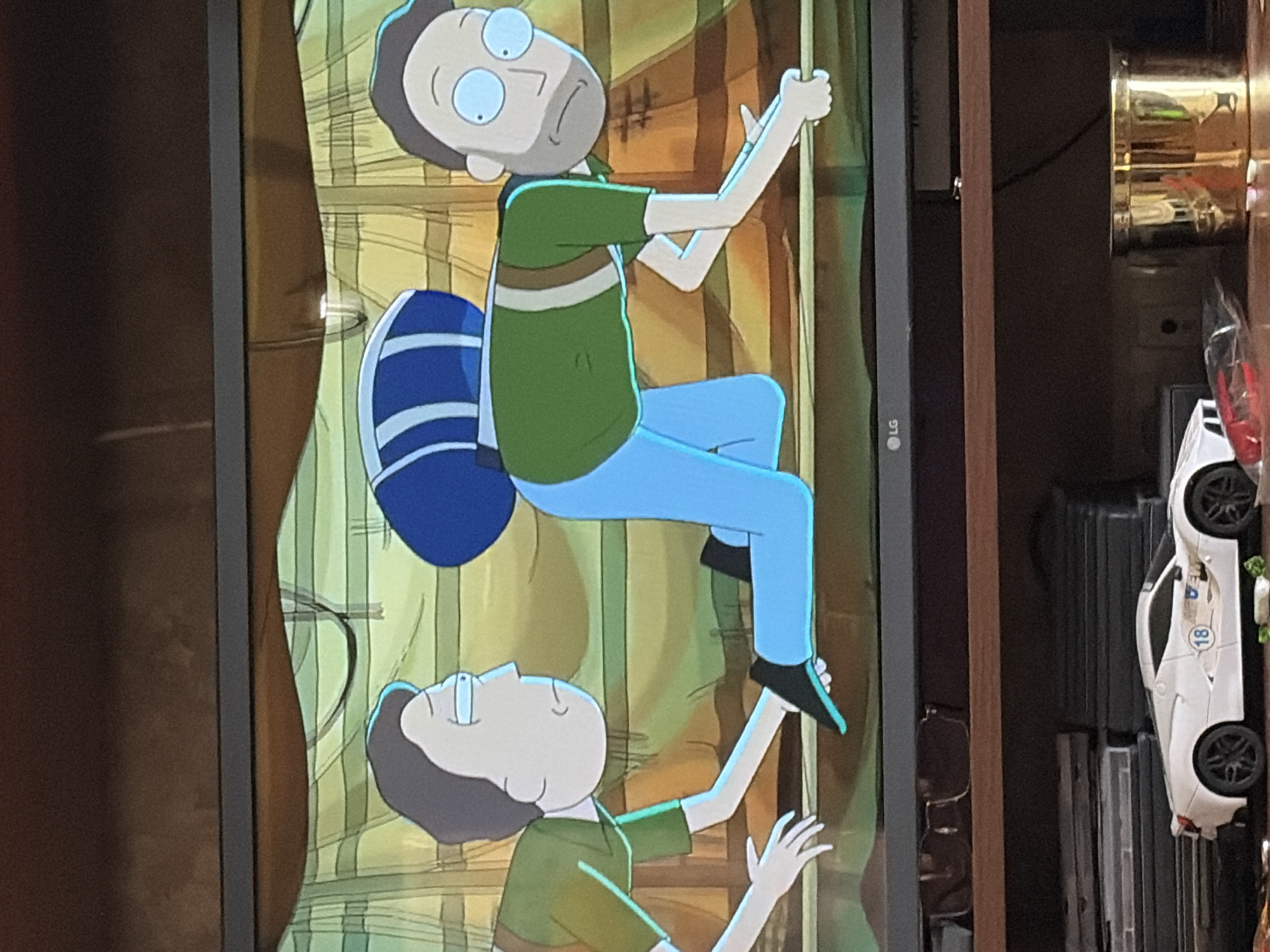The image shows a television screen displaying a scene from the animated show Rick and Morty. The scene depicts two male characters on what appears to be a primitive, wooden structure. The character on the left, resembling Morty Smith, is on his hands and knees, reaching out with his left hand towards the other character. He is wearing a green shirt and light blue pants. The character on the right, resembling Jerry Smith, is also on his hands and knees, with a large blue bag strapped to his back. He is wearing a green shirt with brown straps and light blue pants. His head is turned towards the viewer, and he has a concerned expression. The background of the scene is rendered in muted greens and yellows, with vertical wooden planks visible. Below the television screen, a wooden shelf holds various items, including a white toy car with the number 18 on its side, a pile of DVDs, and a gold-colored container. The television itself has an LG logo on its lower bezel.