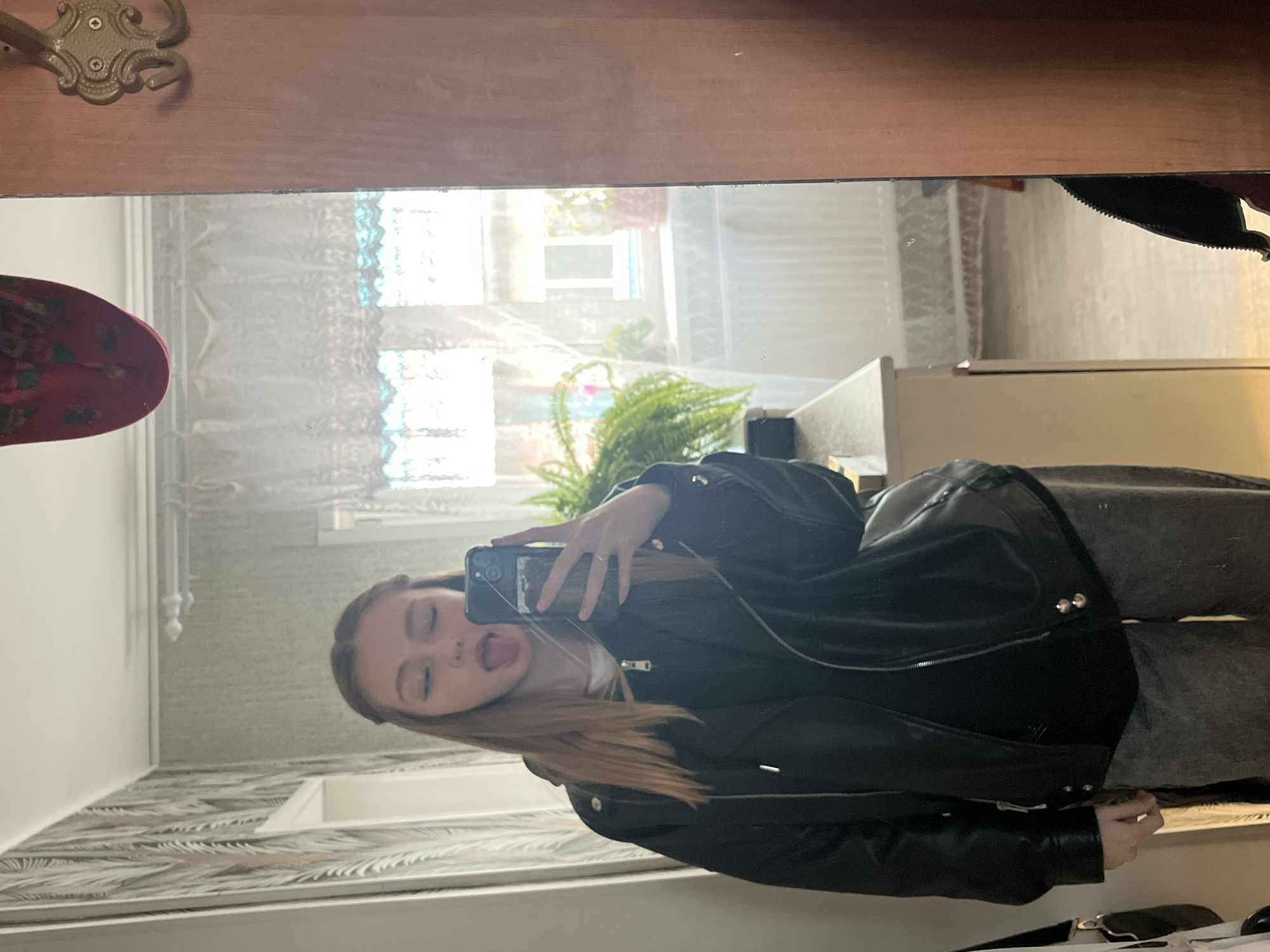 A young woman is taking a selfie in a mirror. She is sticking her tongue out and holding a smartphone. She is wearing a black leather jacket and grey pants. The reflection in the mirror reveals a room interior with wallpaper, a fern plant, and a window with lace curtains. The location appears to be Chaikovsky, Russia, based on the presence of a curtain pattern.