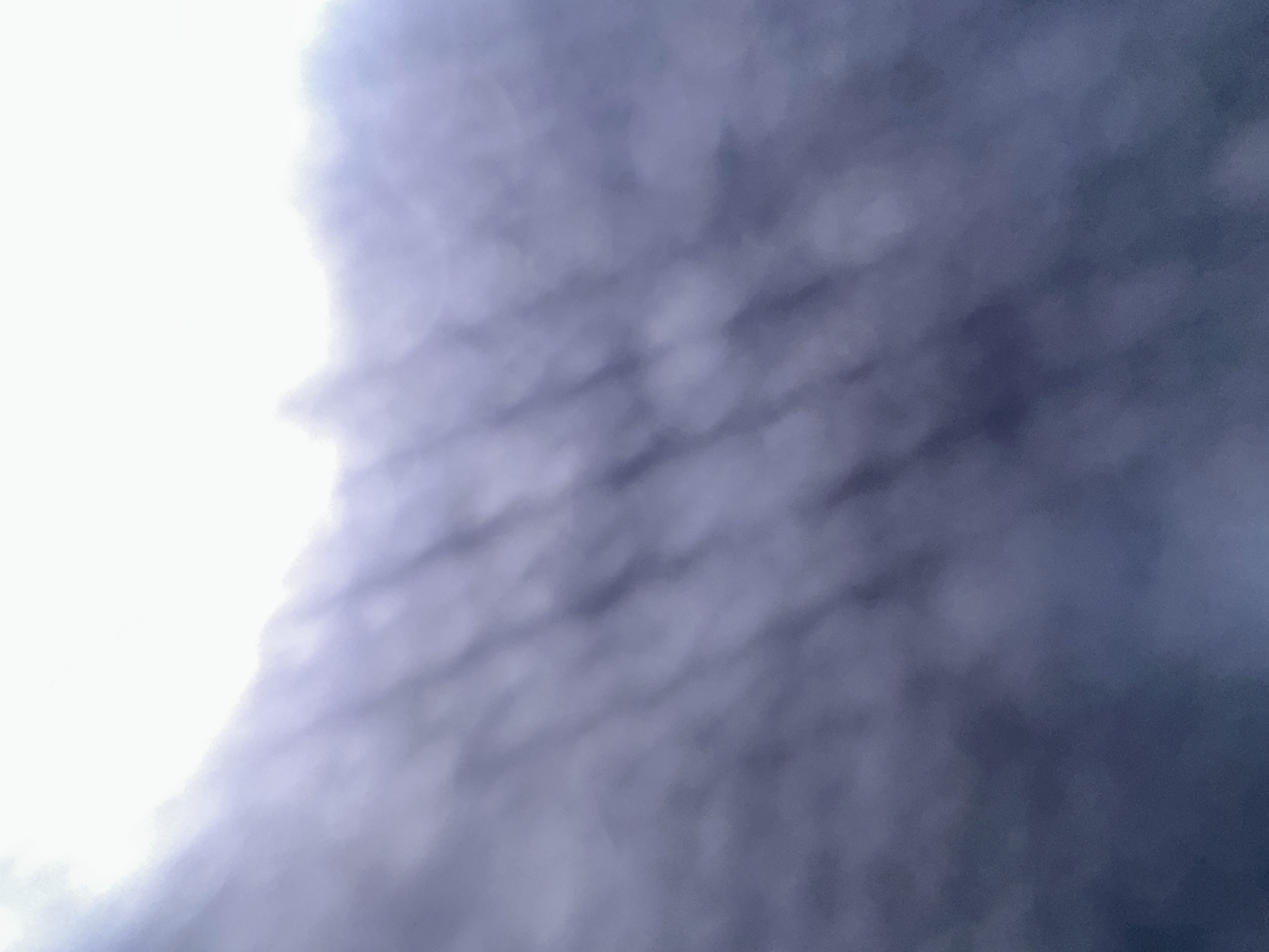 The image depicts a cloudy sky, likely taken from a low angle looking upwards. The left side of the image is dominated by a bright, blown-out area, suggesting a light source such as the sun or a very bright cloud. This bright area extends into the center of the frame, creating a fuzzy, white-to-light-purple gradient. The rest of the image is filled with dark, textured clouds. These clouds appear to have a rippled or layered pattern, with darker, shadowed areas interspersed with lighter, more illuminated sections. The overall impression is of a dramatic or overcast sky. There is no discernible text, people, or specific objects visible. The scene suggests a moment during the day when clouds are prominent, possibly indicating impending rain or a general overcast condition. The location context of Belgrade, Serbia, is noted, but no specific landmarks or visual cues within the image confirm this.