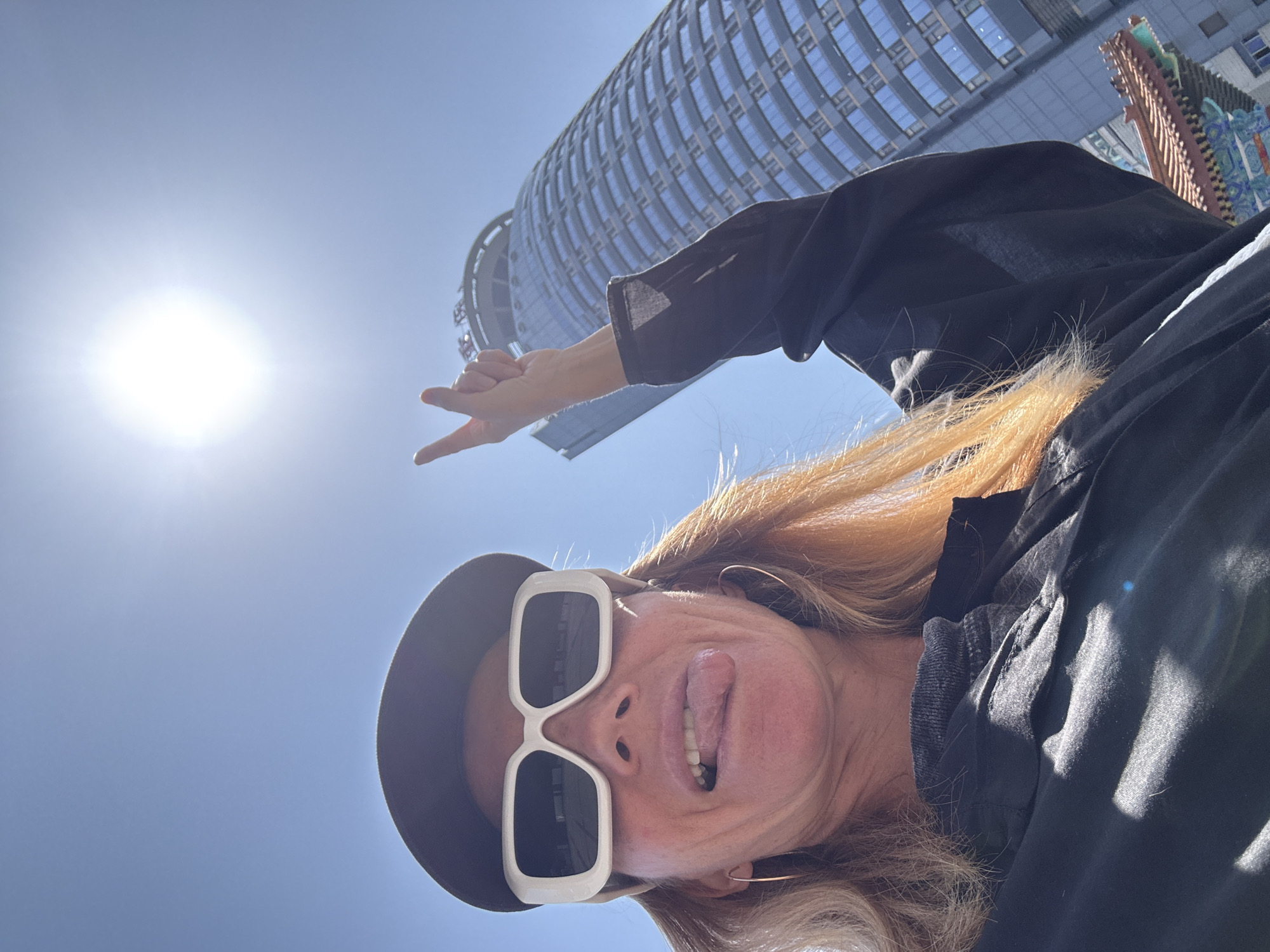 A selfie taken in Yuzhong District, China, features a woman wearing sunglasses and a hat, sticking her tongue out and making a peace sign with her hand. In the background are modern buildings, including a cylindrical tower, under a bright, sunlit sky.