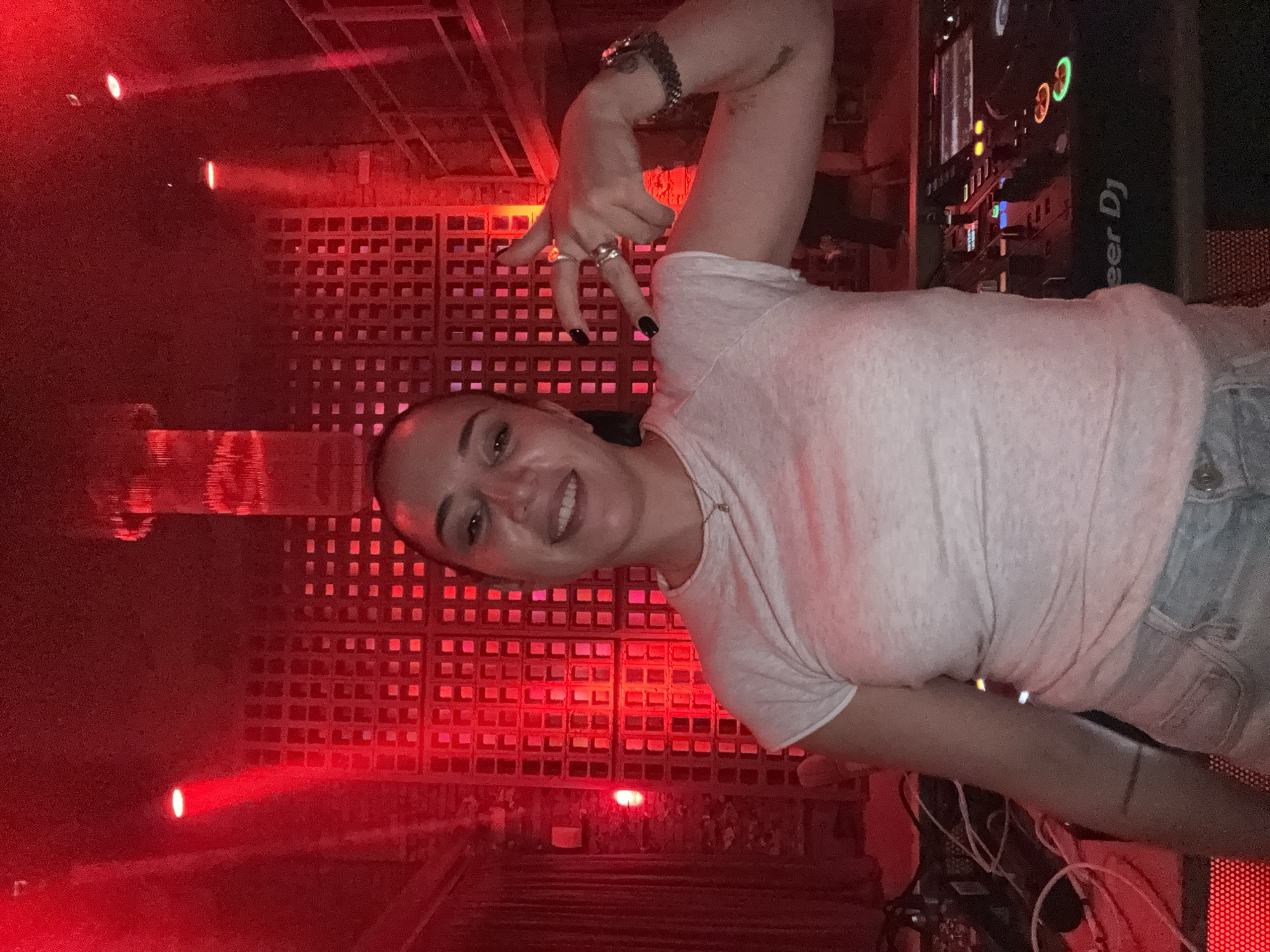 The image shows a woman smiling and making a hand gesture in a room with red lighting. Behind her is a wall with a grid-like pattern. A DJ console labeled "Pioneer DJ" is visible to her right. The location appears to be a nightclub or event venue. The city and country are Moscow, Russia.