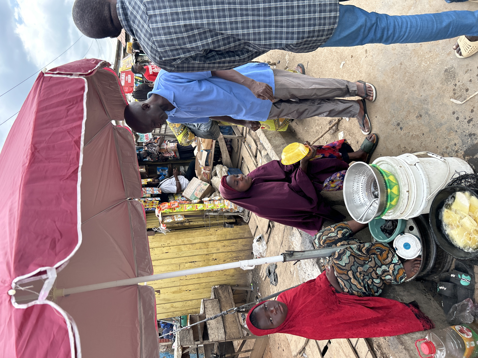 This image depicts an outdoor market scene in the city of Jos, Nigeria. A large, red market umbrella with white scalloped trim is open, providing shade, with its white central pole grounded between two seated individuals. The sky, visible in the upper right, is overcast.
On the left, a young girl wearing a red hijab and a patterned skirt sits barefoot on a low, dark cylindrical object. She looks directly towards the viewer. To her right, a young woman, also seated on a similar low object, wears a deep purple hijab and a floral skirt. She holds a yellow plastic bowl in her left hand and looks towards a standing man. Her right foot, adorned with a green slide sandal, is visible.
A man in a light blue polo shirt and gray trousers stands in front of the woman in purple, facing her. He wears dark flip-flops and is slightly bent forward, his right hand partially open, suggesting an interaction or transaction. To the right, another man is visible from the back, wearing a black and white plaid shirt, blue jeans, and light-colored flip-flops.
The ground is unpaved, appearing as a mix of dirt and concrete. Adjacent to the seated girl, a clear plastic bottle with a red cap, containing a yellow liquid, rests on the ground next to a black sandal. Near the woman in purple, a collection of items includes a stack of white plastic buckets, a green plastic bowl, a metal colander, and a black pot filled with frying yellow food items in oil. A smaller dark pot sits nearby.
The background features a yellow corrugated metal wall or structure. Above this, a red sign with partial white lettering, "IMMANUEL STORES" and a phone number, is visible. Various packaged goods are suspended from strings or displayed on shelves behind the main subjects. Wooden crates and scattered debris are present on the ground. Overhead electrical wires are visible against the sky. Several other individuals can be seen in the mid-ground, indicating a busy public space.