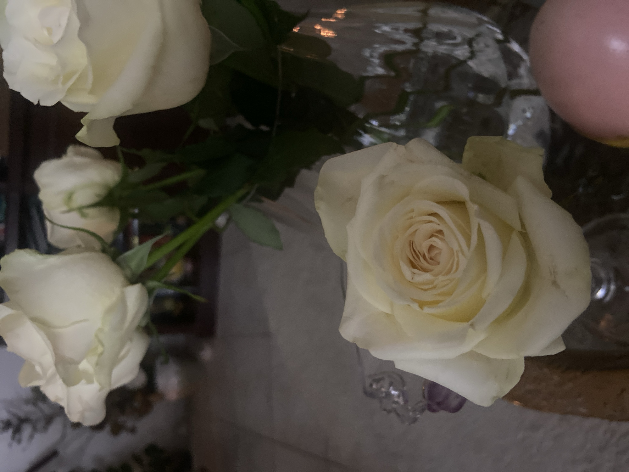 A close-up, low-angle shot focuses on a bouquet of white roses arranged in a clear glass vase. The roses, with their creamy white petals, vary in bloom, with one in the foreground being fully open, revealing its intricate center. The background is dimly lit and blurred, with hints of a textured surface, possibly a table, and indistinct objects, including what appears to be a decorative crystal item and a soft pink spherical object. The overall scene suggests an indoor setting, potentially a home or a florist's shop, but there are no specific visual cues to confirm its location within Deira, United Arab Emirates.