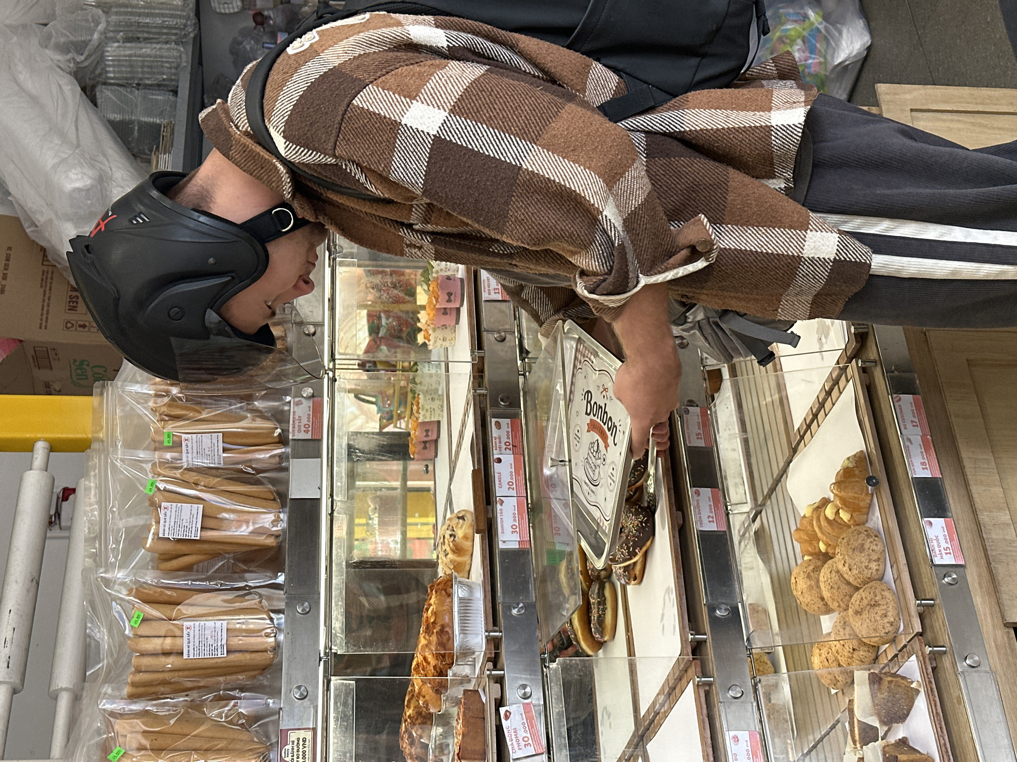A young adult, wearing a black helmet and a brown plaid shirt over dark pants with white stripes, is standing in a bakery in Da Lat, Vietnam. They are holding a silver tray with donuts. The bakery displays a variety of pastries and baked goods on shelves behind glass cases. Packaged breadsticks are visible on an upper shelf. The overall environment suggests a commercial establishment focused on selling baked goods.