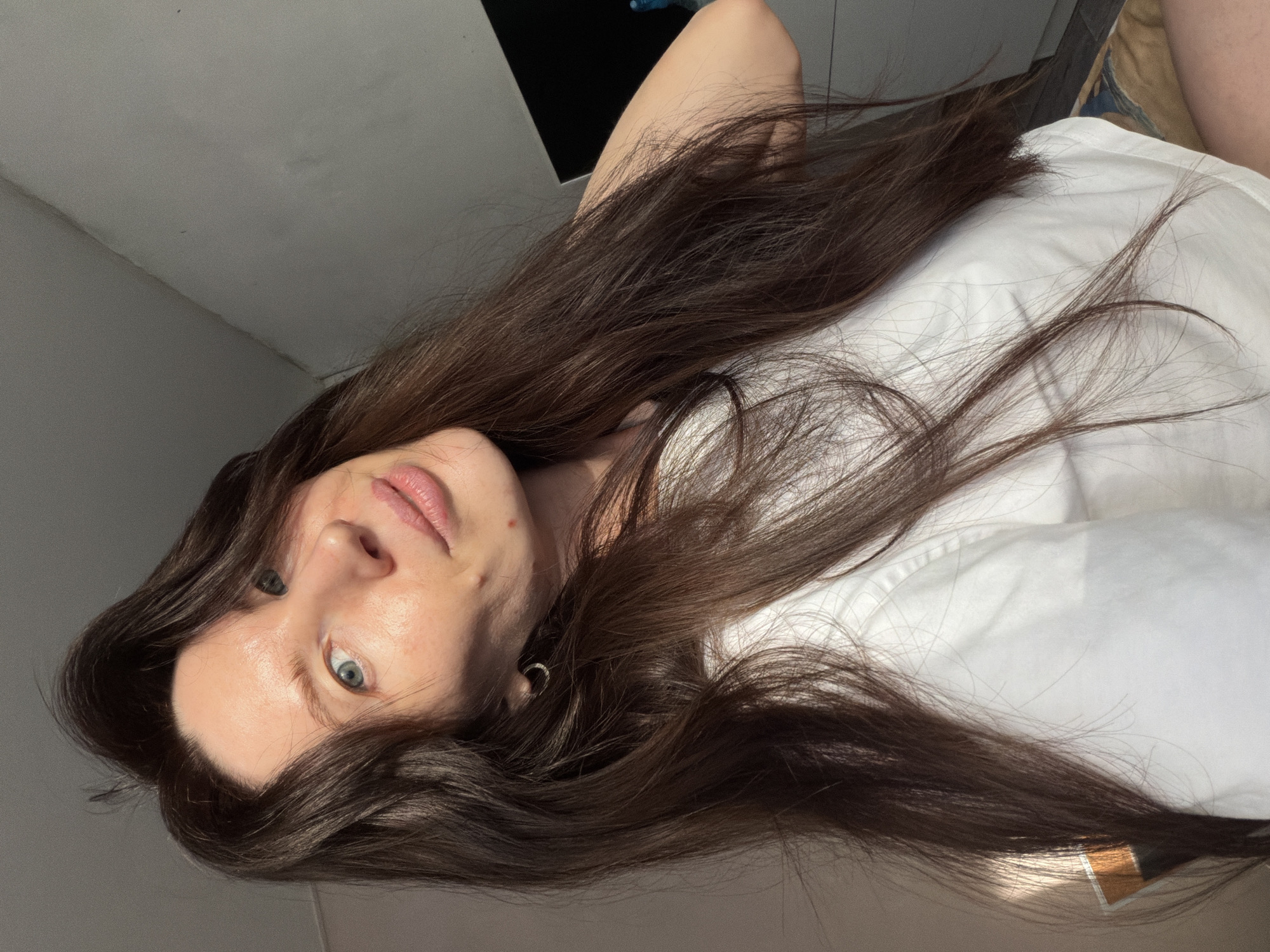 A female individual is depicted in a head-and-chest level self-portrait. She has long, dark brown hair, blue-green eyes, and fair skin, with a faint red mark visible near her mouth and another on her chin. A small silver hoop earring is present in her visible left ear. She is wearing a plain white t-shirt. Her head is tilted slightly, and she maintains direct eye contact with the camera, exhibiting a subtle, closed-mouth smile. The scene is an indoor residential environment. The background features a light grey wall in the upper left, showing a subtle texture and a ceiling junction. The upper right background contains a darker area with a partial view of a dark rectangular object. In the lower right, patterned fabric, likely part of bedding or upholstery, is visible. Natural light illuminates the subject from the left side. No specific identifiers for the city of Perm, Russia, or any precise location within it, are discernible from the visual information presented in the image.
