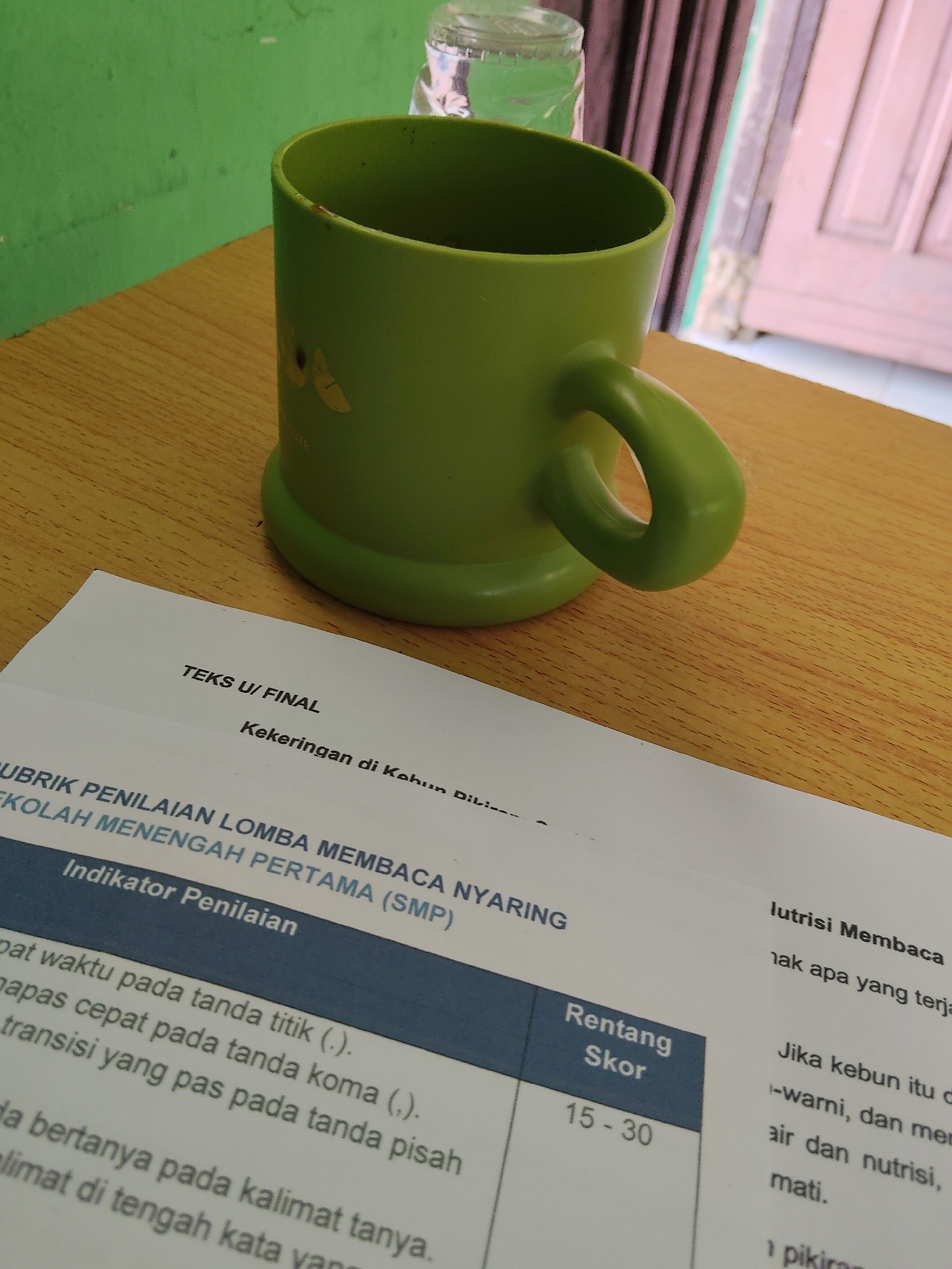 A green mug sits on a wooden table next to a document. The document is titled "TEKS U/ FINAL" and contains text related to "RUBRIK PENILAIAN LOMBA MEMBACA NYARING SEKOLAH MENENGAH PERTAMA (SMP)". The mug is slightly off-center to the left, with its handle facing right. The document is positioned in front of the mug, angled slightly towards the viewer. In the background, a green wall is visible, with a blurry window or door on the right. The lighting suggests an indoor setting, possibly during the day. There are no people visible in the image.