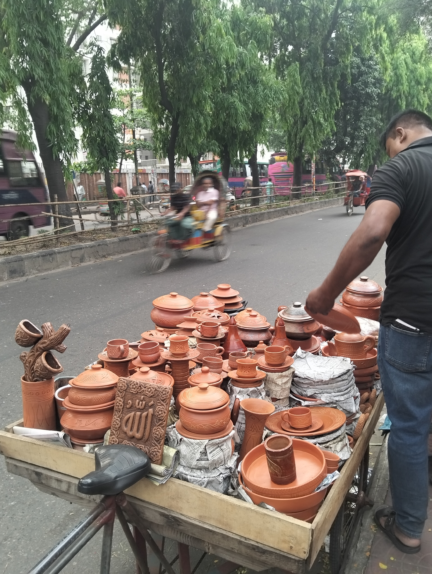 An eye-level, wide shot depicts a street scene in Dhaka, Bangladesh, featuring a vendor and a bicycle rickshaw stall laden with terracotta pottery. The road, visible in the background, shows light vehicular traffic, including a passing bus on the left and a three-wheeled rickshaw traveling to the right. Behind the road, a row of tall, green trees lines the edge of the sidewalk.
In the foreground, a man is seen from the side, standing next to the cart. He is wearing a black shirt and blue jeans and appears to be handling one of the terracotta plates. The cart itself is crammed with a variety of terracotta items, including pots, cups, plates, and decorative pieces, some wrapped in newspaper for protection. A bicycle seat is visible, indicating the cart's mobility. The terracotta items are the focal point of the image. The environment conveys an urban setting with bustling street activity.