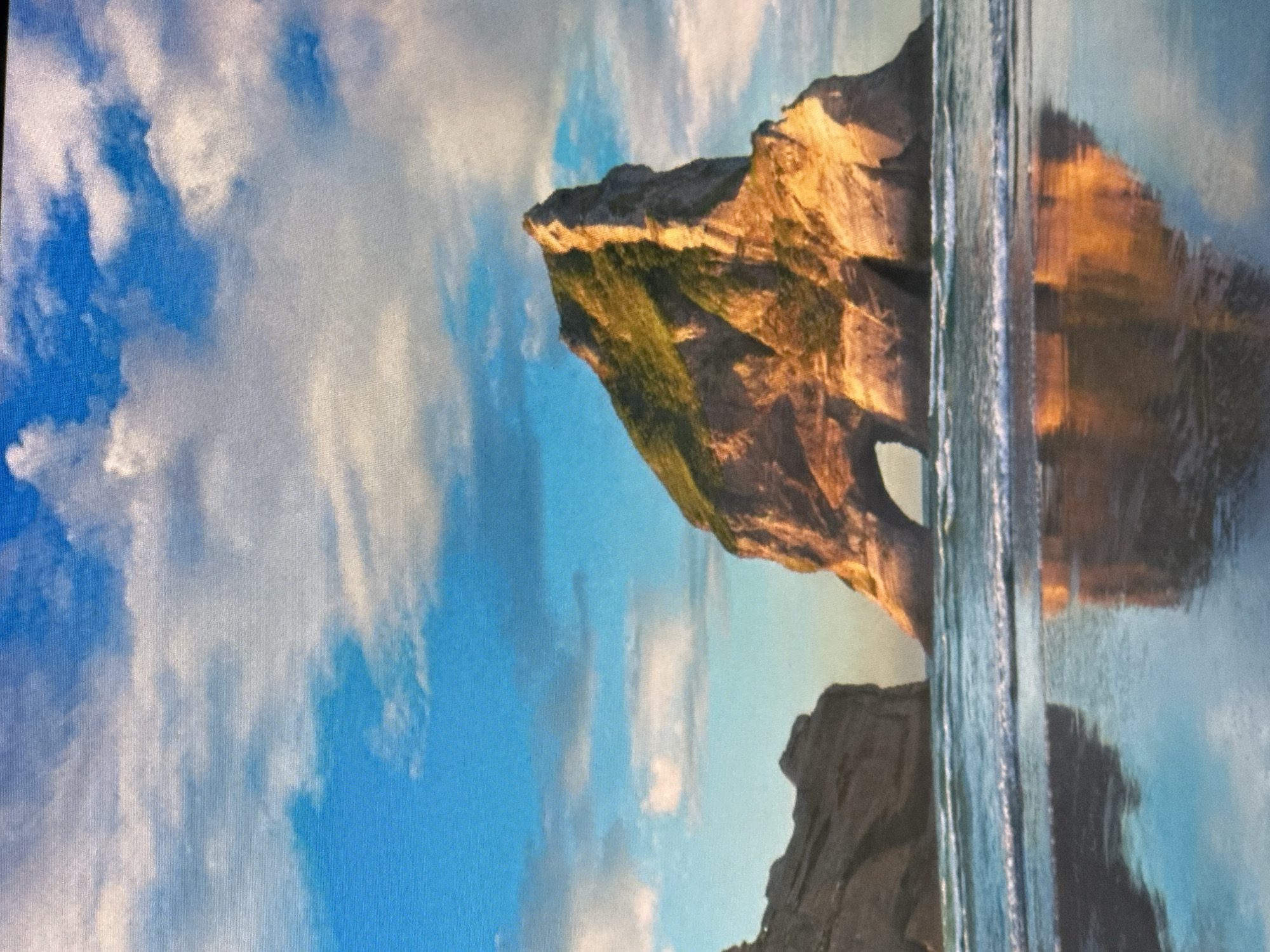 The image depicts a dramatic coastal landscape with a rock formation featuring a natural archway. The rock is illuminated by warm sunlight, highlighting its textured surface and the green vegetation growing on its slopes. The scene includes a body of water, likely the sea, with gentle waves reflecting the sky and the rock formation. The sky is filled with fluffy white clouds against a clear blue backdrop. The reflection in the water is clear, mirroring the rock and sky. Two rock formations are visible, one larger with the arch and a smaller one in the foreground, partially submerged in water. The overall mood is serene and majestic, evoking a sense of natural beauty and tranquility. The time of day appears to be either late afternoon or early morning, given the warm golden light. There is no visible text in the image.