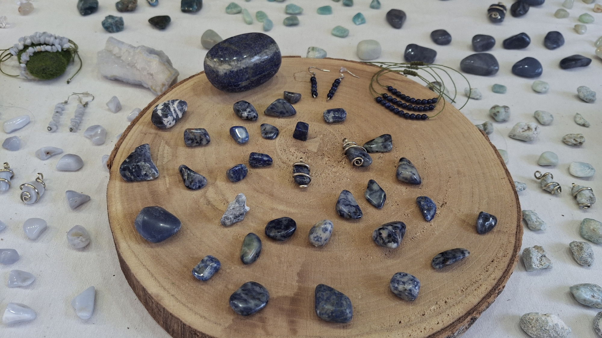 The image shows a collection of natural stones, jewelry, and a wooden slice displayed on a white tablecloth. The stones vary in size, shape, and color, with a focus on blue and grey hues, suggesting they might be lapis lazuli, sodalite, or similar minerals. Some stones are polished and smooth, while others are rough and crystalline.
Several pieces of jewelry are visible, including a pair of dangling earrings with small blue beads and a necklace or bracelet made of similar beads. There are also pendants wrapped in wire. A green mossy sphere with some stones attached is on the left side of the image, possibly part of a decorative arrangement.
The stones are scattered around a prominent, circular wooden slice that serves as a central display piece. The wood grain is clearly visible, indicating it's a cross-section of a tree trunk.
The overall arrangement suggests a market stall or an artisan's display, likely for sale. The context provided about the location being in La Fresneda / la Freixneda, Spain, implies this is an outdoor or indoor market in that specific Spanish town. No people are visible in the image.