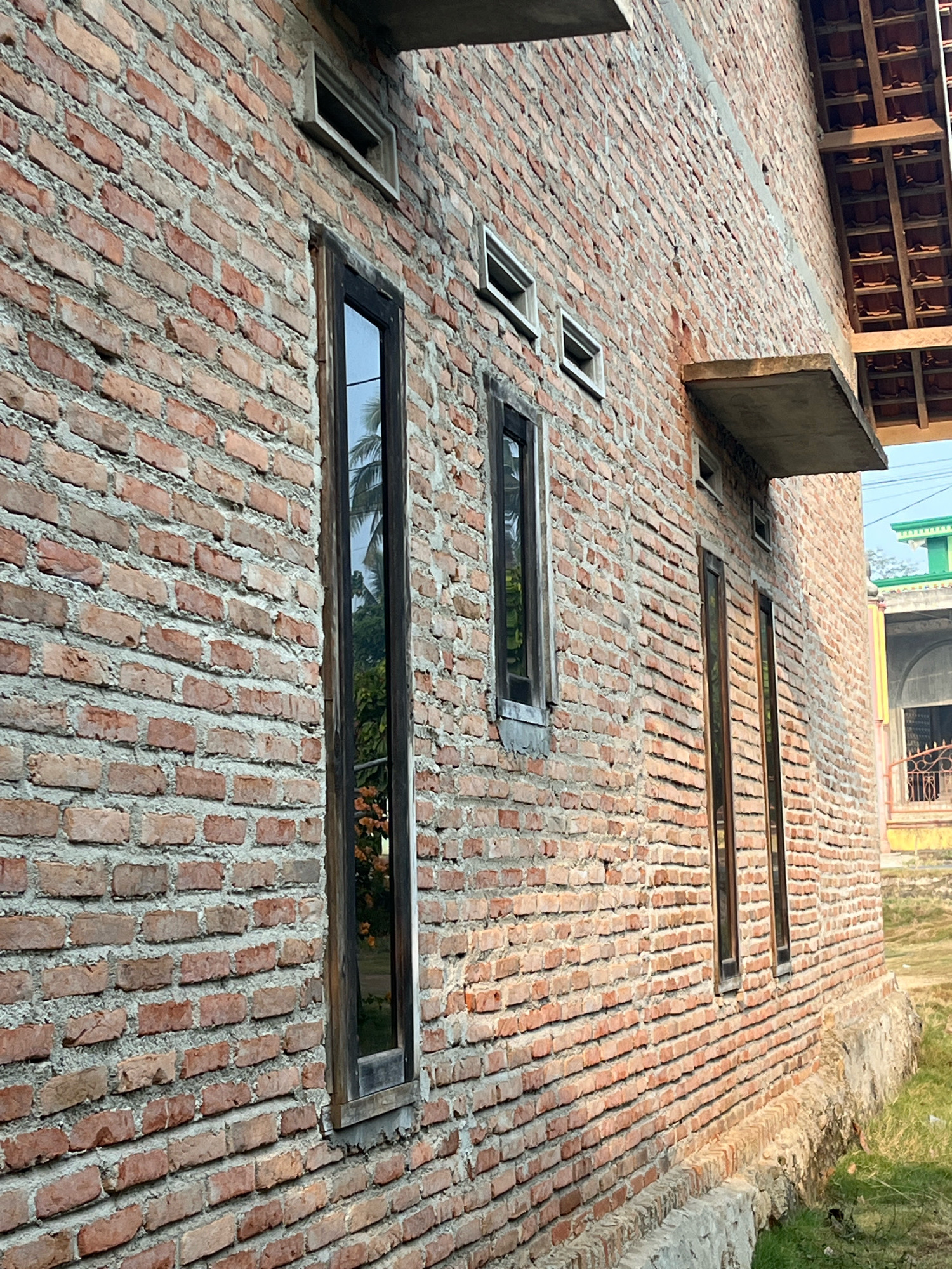 An exterior, eye-level, medium shot shows a brick building in Kebumen, Indonesia. The wall is constructed of reddish-brown bricks laid in a running bond pattern with gray mortar. There are several tall, narrow, dark-framed windows with reflective glass spaced along the wall. The windows reflect palm trees and foliage. Small rectangular vents are present above some of the windows. The roofline, with terracotta tiles and wooden beams, is visible at the top right of the image, as is a concrete overhang. In the background, a portion of another building with a green roof and a decorative metal gate is visible. The ground in front of the wall is covered with sparse green grass and has a concrete foundation.