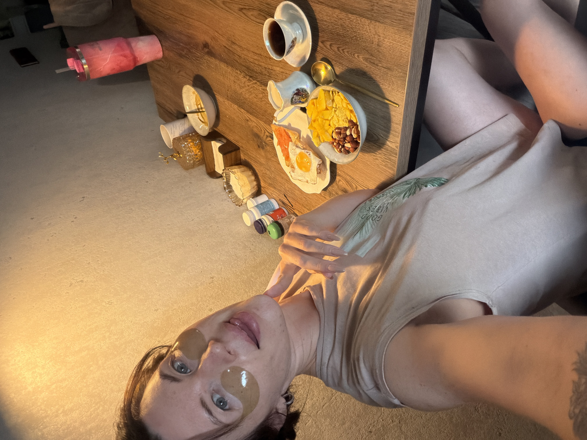 A woman with eye patches is taking a selfie at a wooden table with breakfast. She is wearing a light-colored t-shirt. On the table are a bowl of cereal with fruit and nuts, a plate with a fried egg and toast, a creamer pitcher, and a cup of coffee. A pink tumbler with a straw is in the background, along with some jars and containers. The scene appears to be indoors, likely in a home or apartment, with a textured wall behind the woman. There are no visible landmarks to indicate a specific location in Perm, Russia.