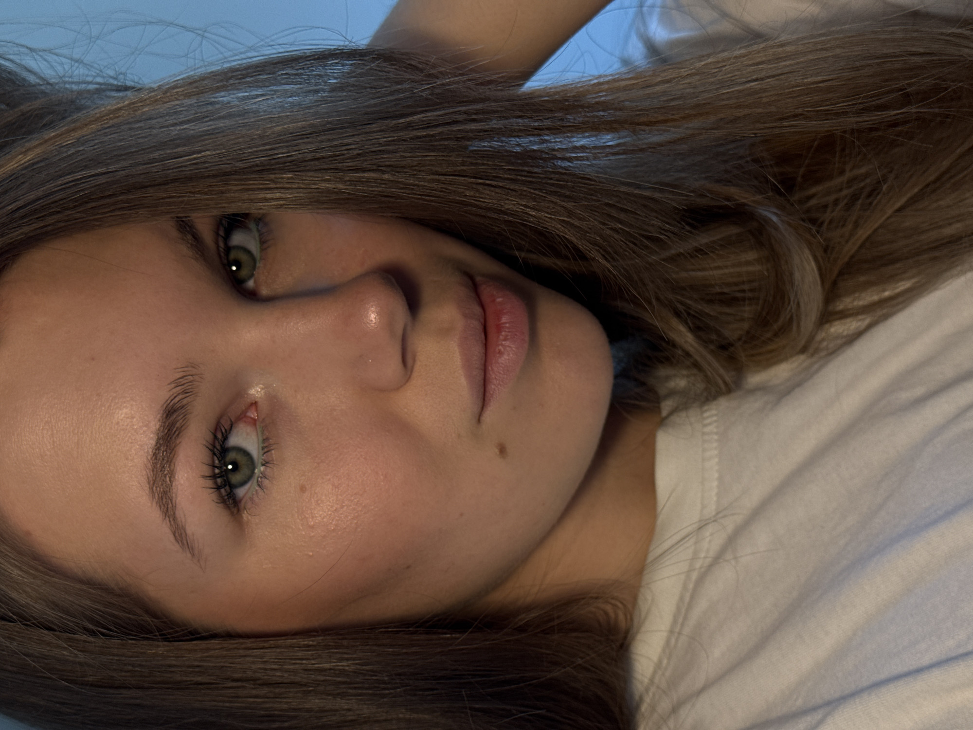 A close-up, portrait-style image shows a young woman with long, brown hair, lying down. She has fair skin, green eyes, and full lips. She is wearing a white t-shirt. There is no visible information to determine if the image was taken in the city of Vozdovac Urban Municipality, Serbia.